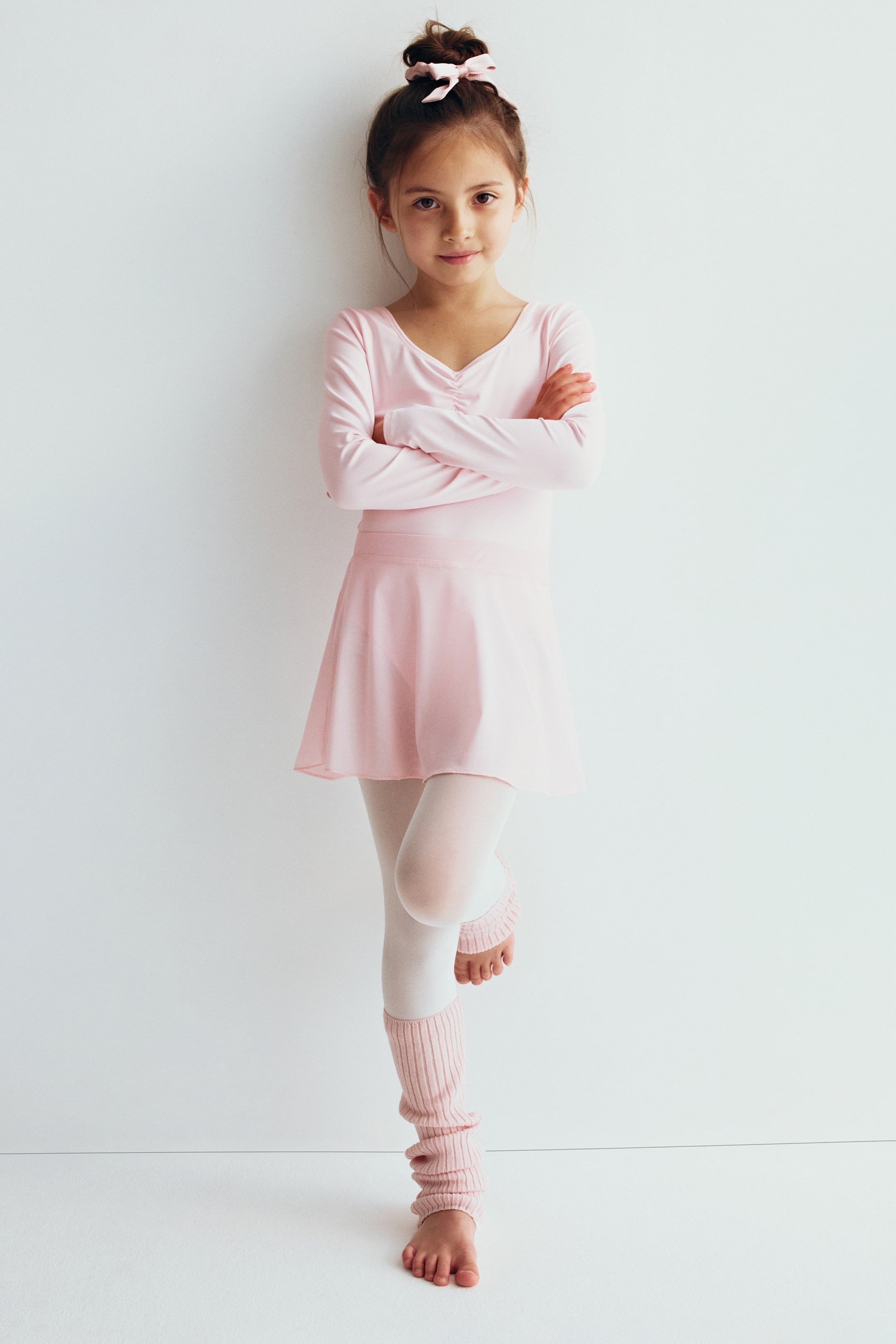 Voir une image plus grande: Une robe de ballet à manches longues rose pâle présente un col en V froncé et une jupe évasée légère intégrée, vue de face. La tenue est stylisée avec des collants blancs et des chauffe-jambes roses côtelés assortis.