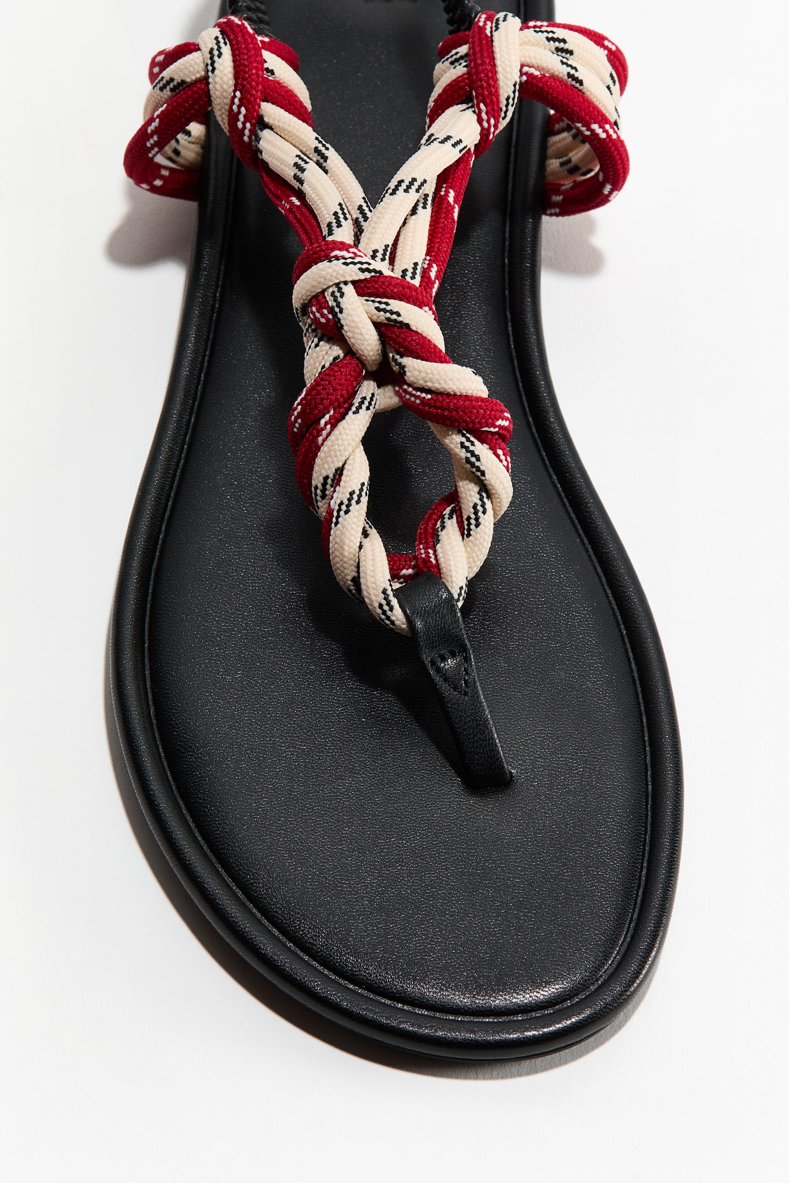 A sandal featuring an almond toe and a narrow black toe post. Its foot straps are crafted from knotted red, beige, and black laces, with the product being presented from a front-facing angle, highlighting the toe area.