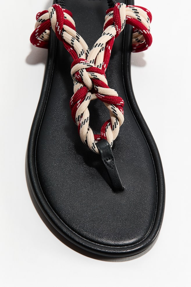 A sandal featuring an almond toe and a narrow black toe post. Its foot straps are crafted from knotted red, beige, and black laces, with the product being presented from a front-facing angle, highlighting the toe area. - 1