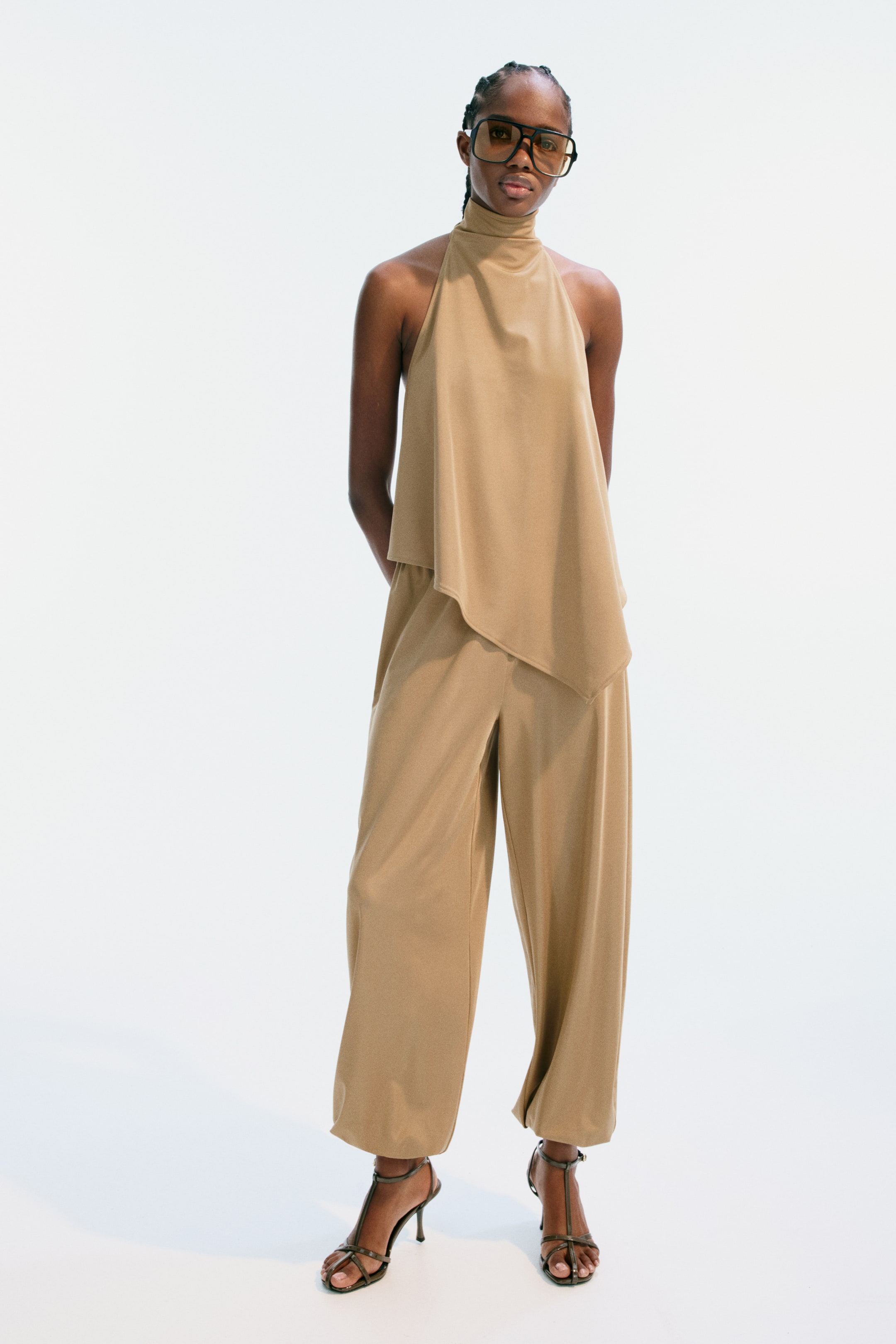 Ampliar la imagen: Un conjunto coordinado en color tostado presenta un top de cuello halter con escote alto y un dobladillo asimétrico, combinado con pantalones anchos a juego que se acumulan en los tobillos. Sandalias de tacón alto con tira en T oscura completan el look.