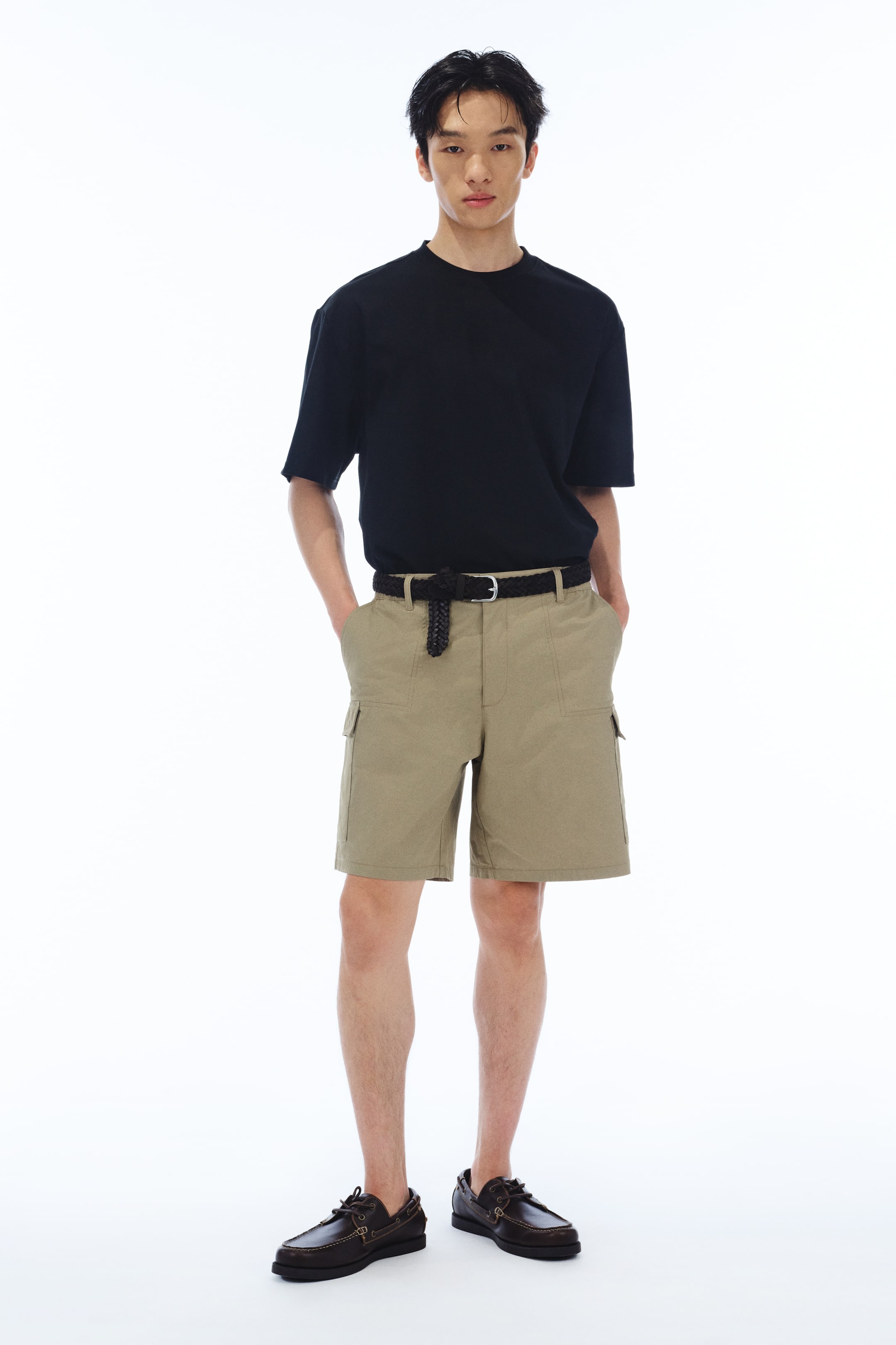 拡大画像を表示: モデルが着用しているのは、黒のTシャツです。ヘビーウェイトのジャージーコットンブレンドで作られており、丸いリブトリムのネックライン、ドロップショルダー、ストレートカットの裾が特徴です。ゆったりとしたシルエットを提供するルーズフィットで、カーキ色のショートパンツにタックインされています。ショートパンツは編み込みベルトで固定されています。