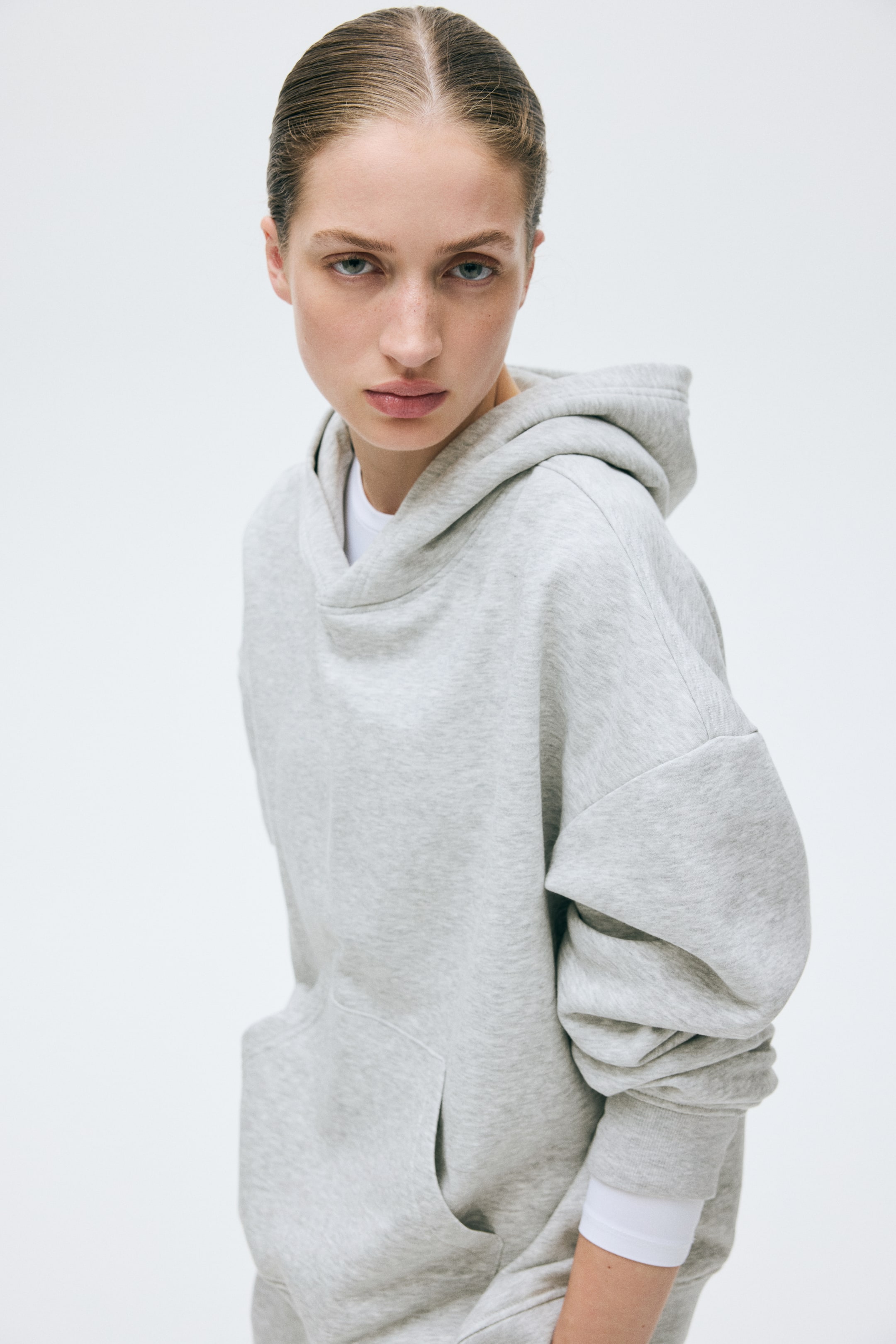 View larger image: Presenting a light gray oversized hoodie, featuring a relaxed hood, dropped shoulder seams, and a front kangaroo pocket, with a white long-sleeve shirt layered underneath the cuffs and neck opening.