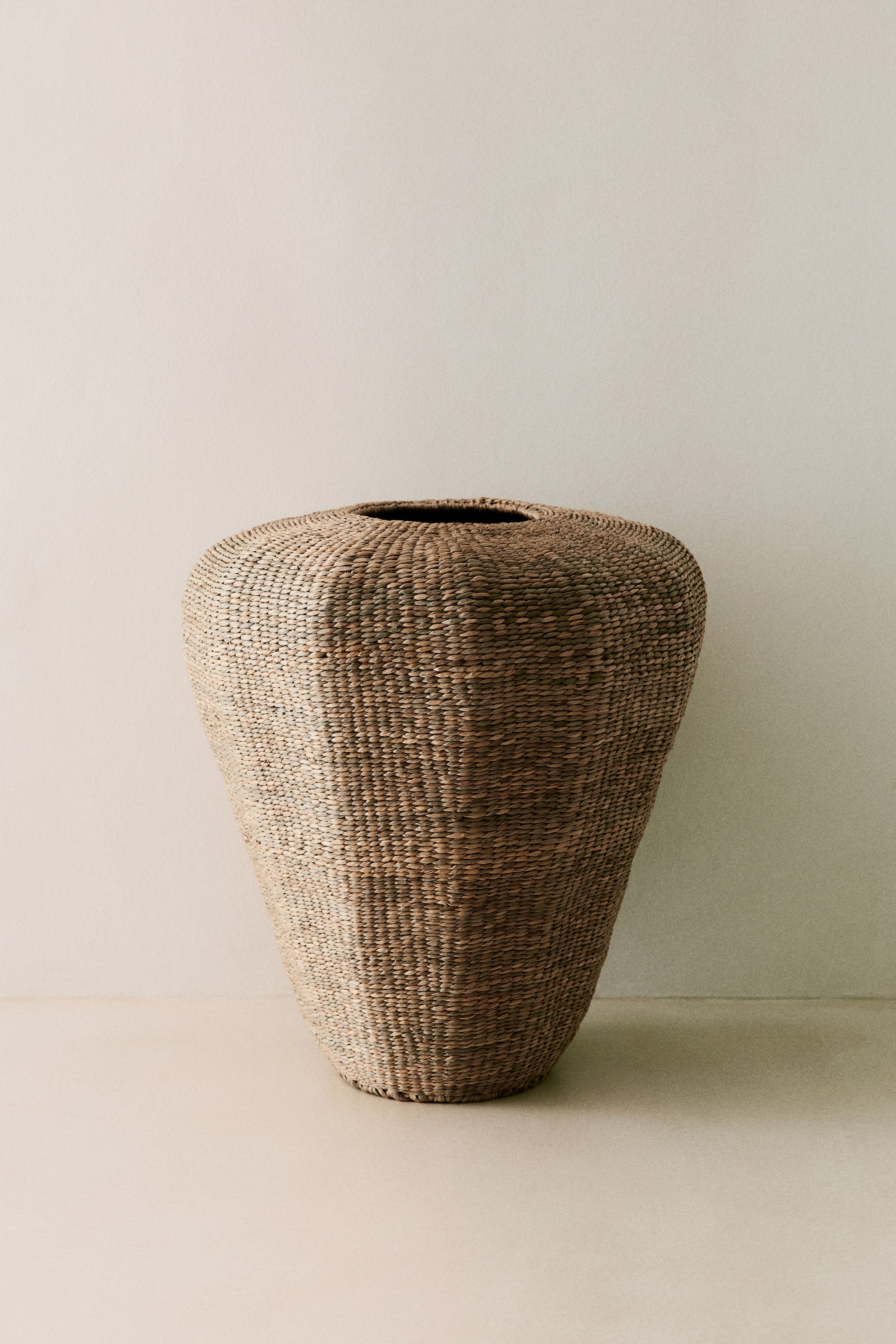 View larger image: A large woven basket stands upright, showcasing a wide, rounded upper body with a narrow opening. Its tightly textured natural fiber weave tapers downwards to a slightly flared base.