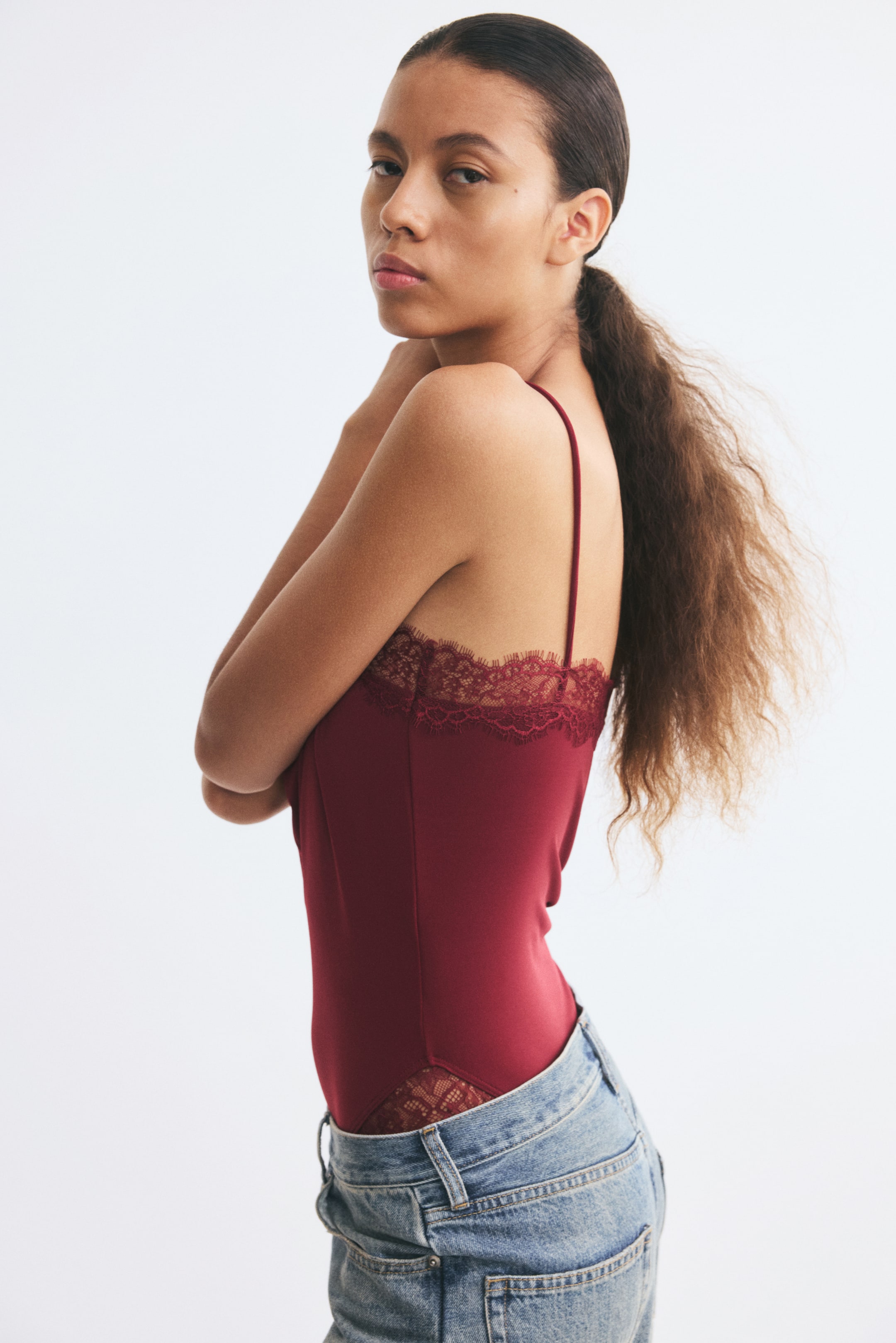 View larger image: A burgundy jersey bodysuit with thin spaghetti straps and delicate lace trim along the straight neckline and leg openings is worn by the model. The bodysuit is styled with low-rise, light-wash blue jeans.