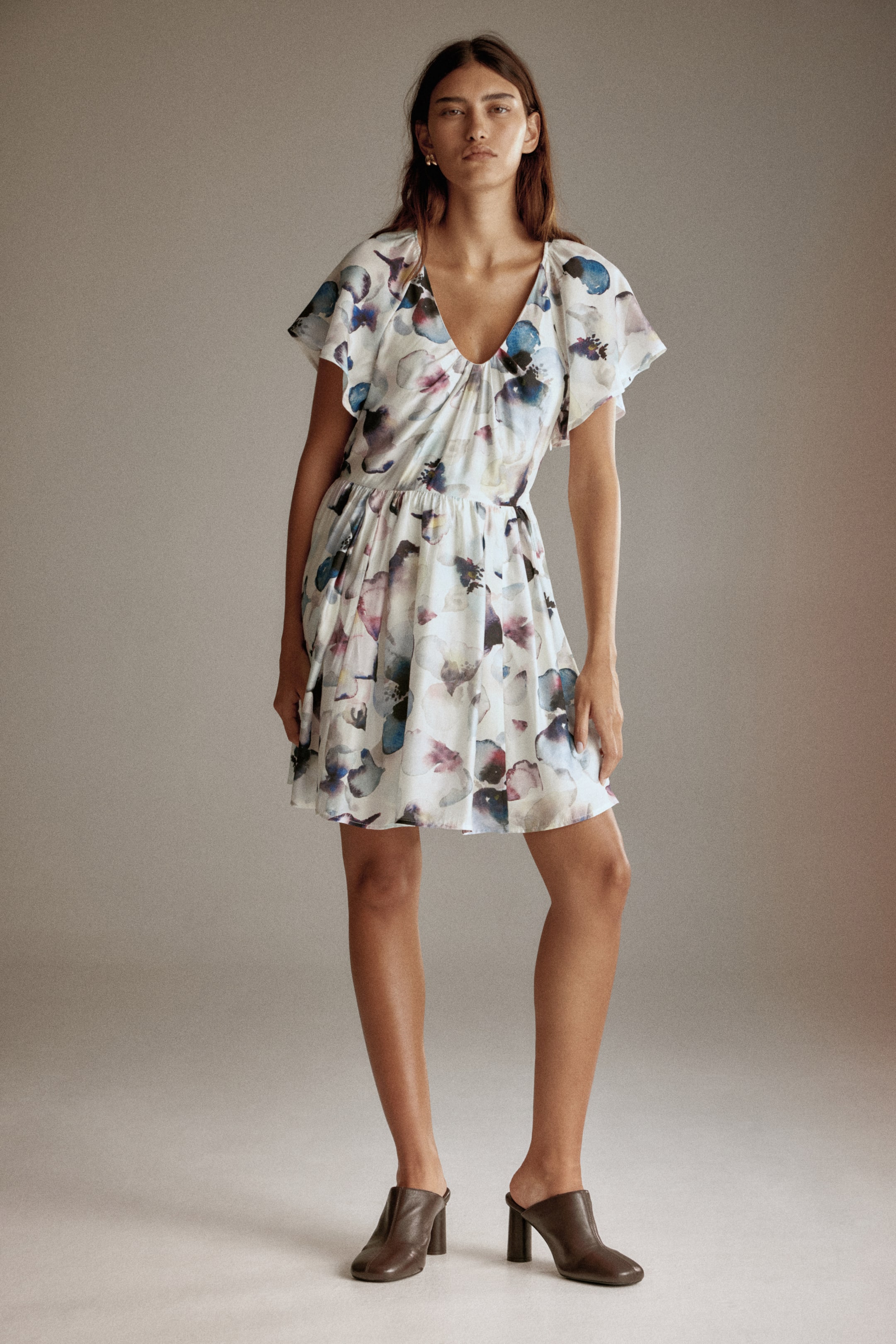 Xem ảnh lớn hơn: Một chiếc váy trắng mặt trước có họa tiết hoa vẽ màu nước với các sắc thái của xanh lam, tím và xanh lá cây. Nó được thiết kế với cổ chữ V xếp ly, tay ngắn xòe nhẹ và eo thun, bắt đầu nở rộng thành chân váy ngắn xòe. Chiếc váy được mặc cùng với giày mule da nâu.