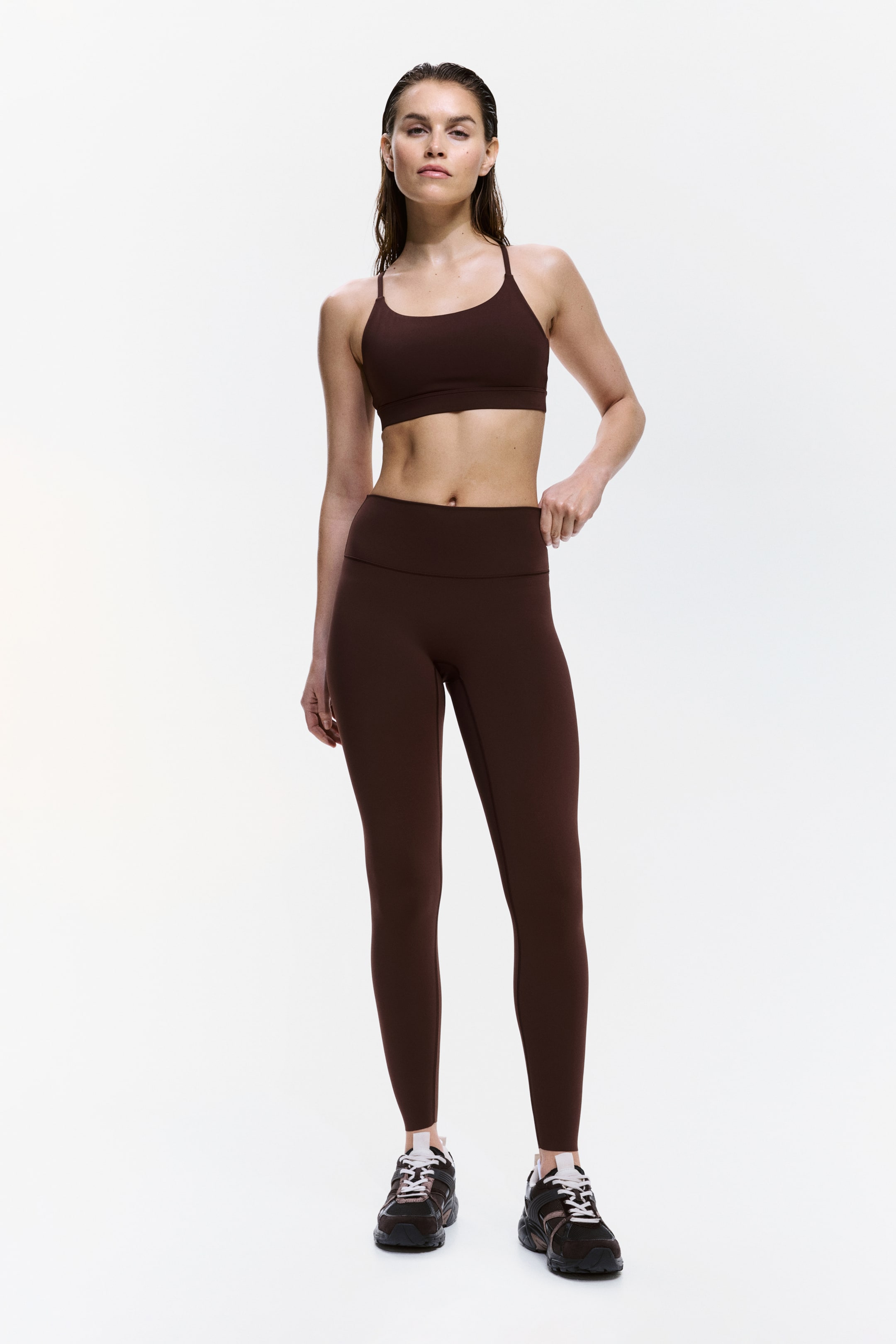 Grotere afbeelding bekijken: Een donkerbruin activewear set wordt gepresenteerd met de voorkant naar voren, met een sportbeha met ronde hals en spaghettibandjes en bijpassende hoog getailleerde, lange leggings, beide met een nauwsluitend ontwerp.