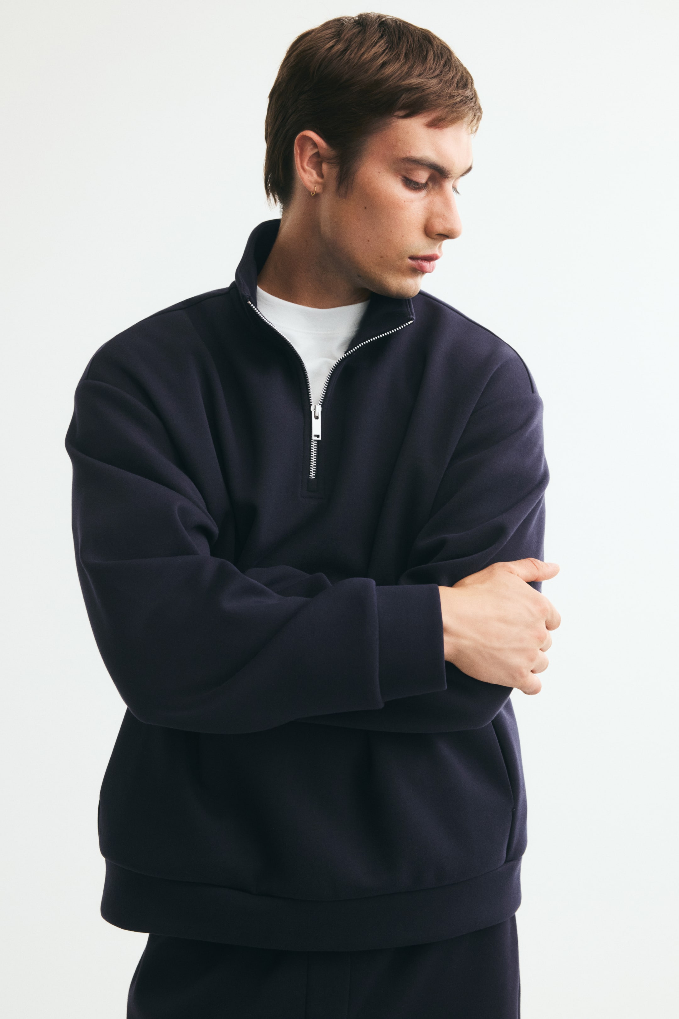 Voir une image plus grande: Un chandail demi-zip à coupe décontractée, bleu marine foncé, est porté par le mannequin, qui est debout avec les bras croisés. Le chandail présente un col montant avec une fermeture éclair argentée partiellement ouverte laissant voir un t-shirt blanc superposé en dessous, des épaules tombantes et des poignets côtelés.