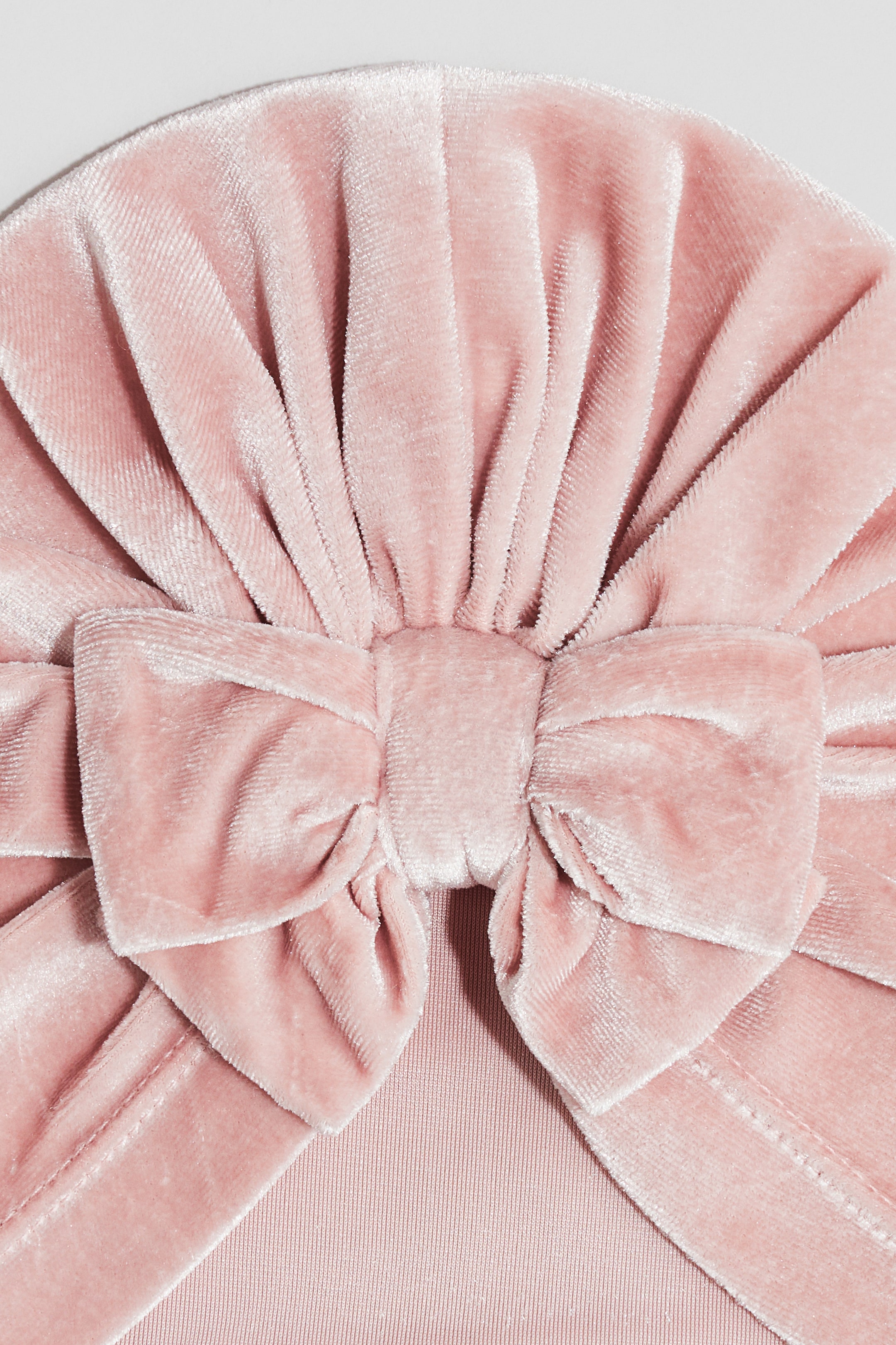 Ingrandisci l'immagine: Un turbante di velluto rosa mostra un grande fiocco centrato sulla parte anteriore. Il tessuto di velluto forma pieghe radianti attorno al fiocco, e una base a maglia a coste è visibile sotto.