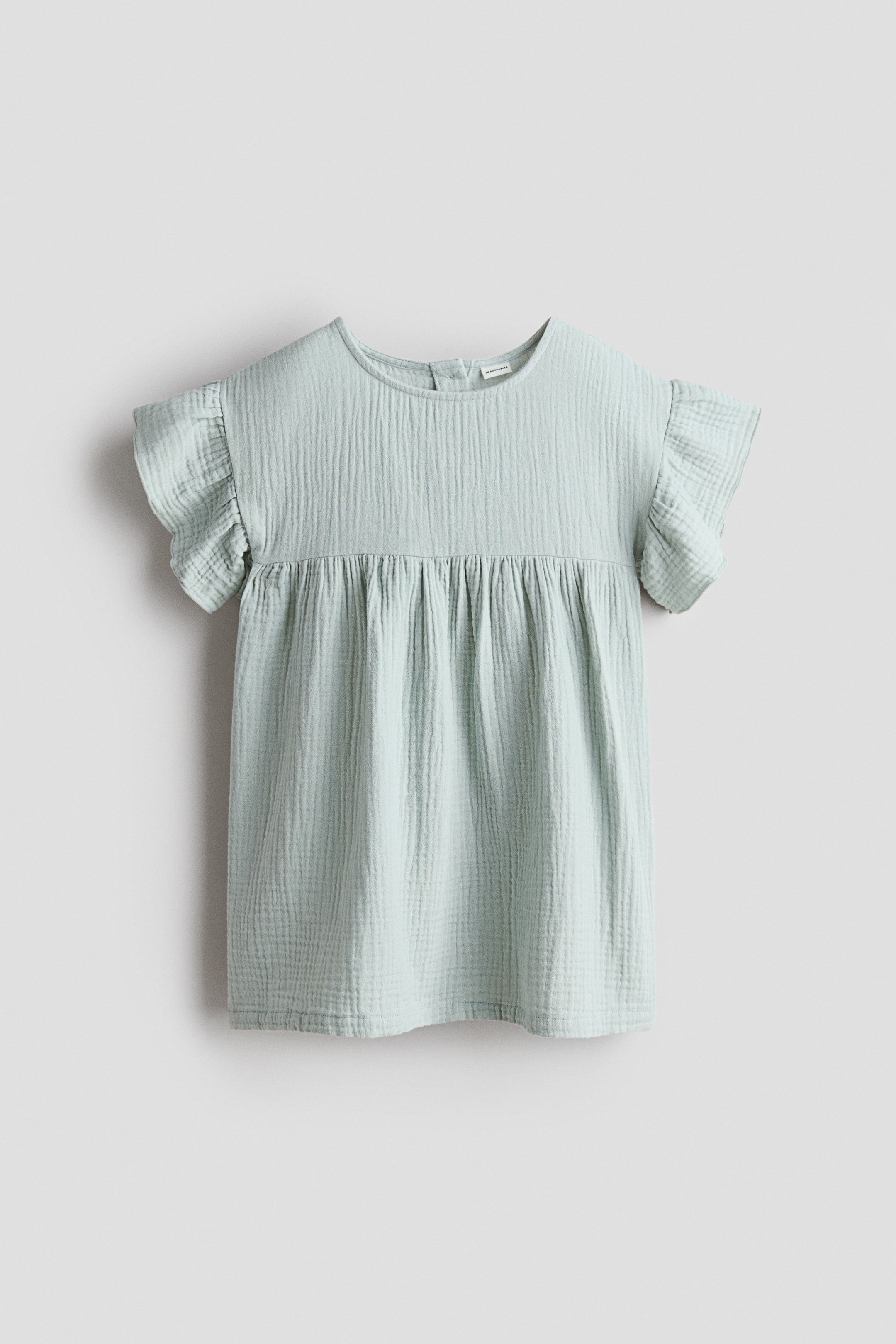 Büyük resmi görüntüle: H&M Adorables elbisenin ön görünümü sunuluyor: Açık yeşil muslin kumaştan yapılmış, yuvarlak yakalı, düşük omuzlu ve kısa kanatlı kollu bir elbise. Göğüs altındaki büzgülü dikiş, genişleyen bir eteğe yol açıyor ve yakanın arkasında küçük, yuvarlak düğmeler görülüyor.