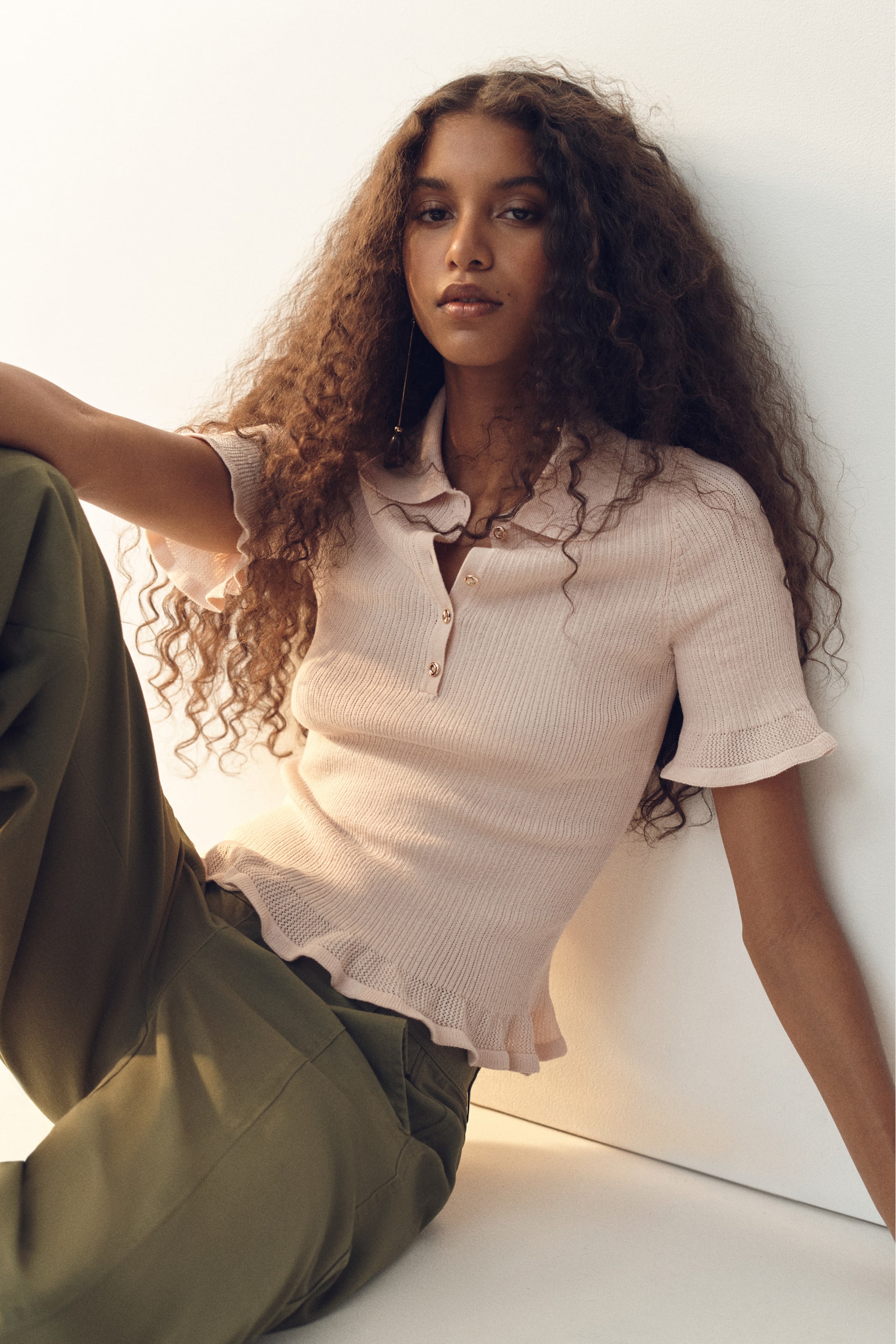 View larger image: The model is wearing a fitted top crafted from soft rib knit fabric. It features a collar, buttons at the top, short sleeves, and frill trims at the cuffs and hem. The top is paired with olive green pants.