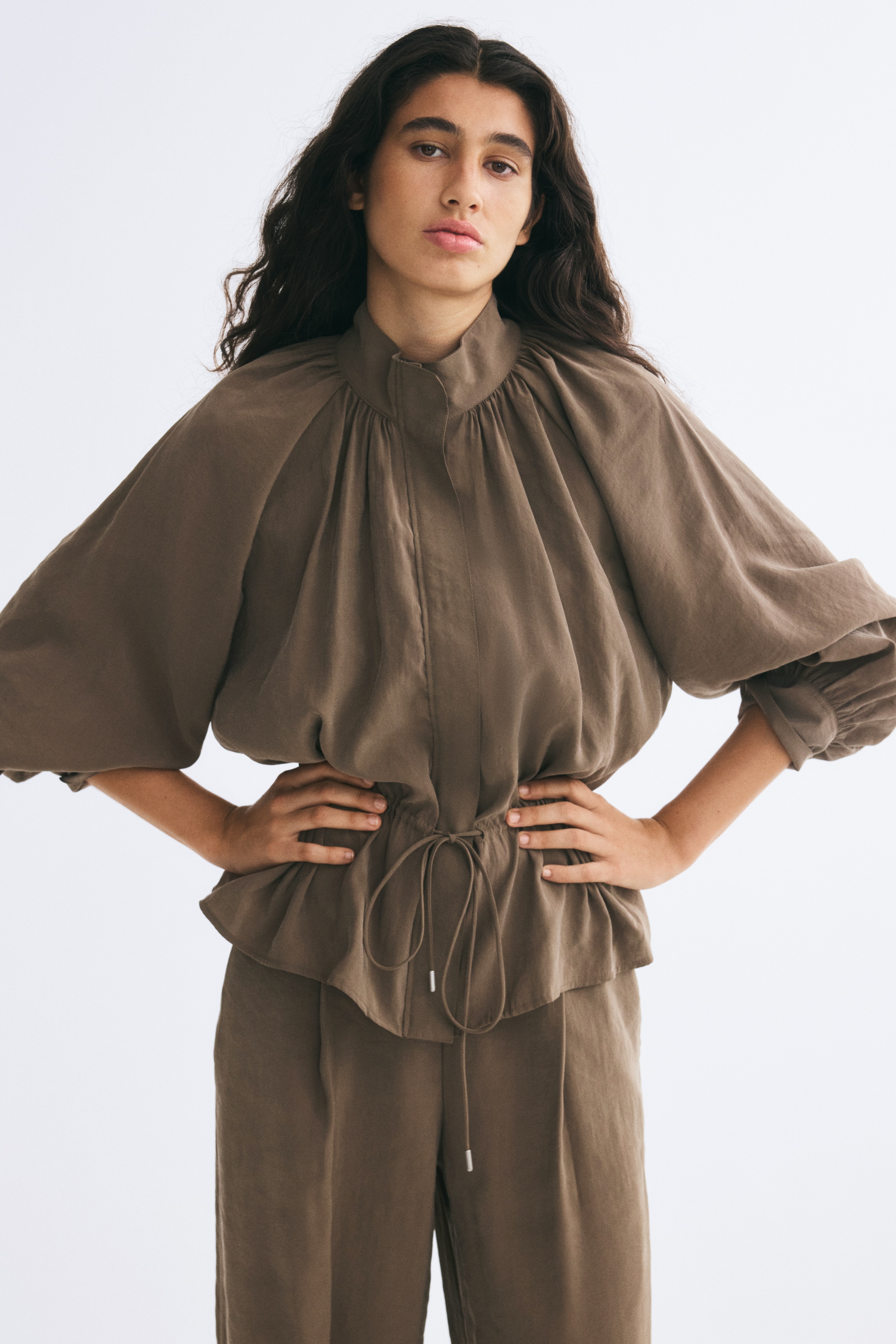 A brown modal-blend jacket is shown from the front, featuring a high stand collar, a concealed front placket, and gathered raglan balloon sleeves with wide cuffs. The jacket is cinched at the waist with an adjustable drawstring and tied cords, creating a gathered effect that flares out into a peplum hem.