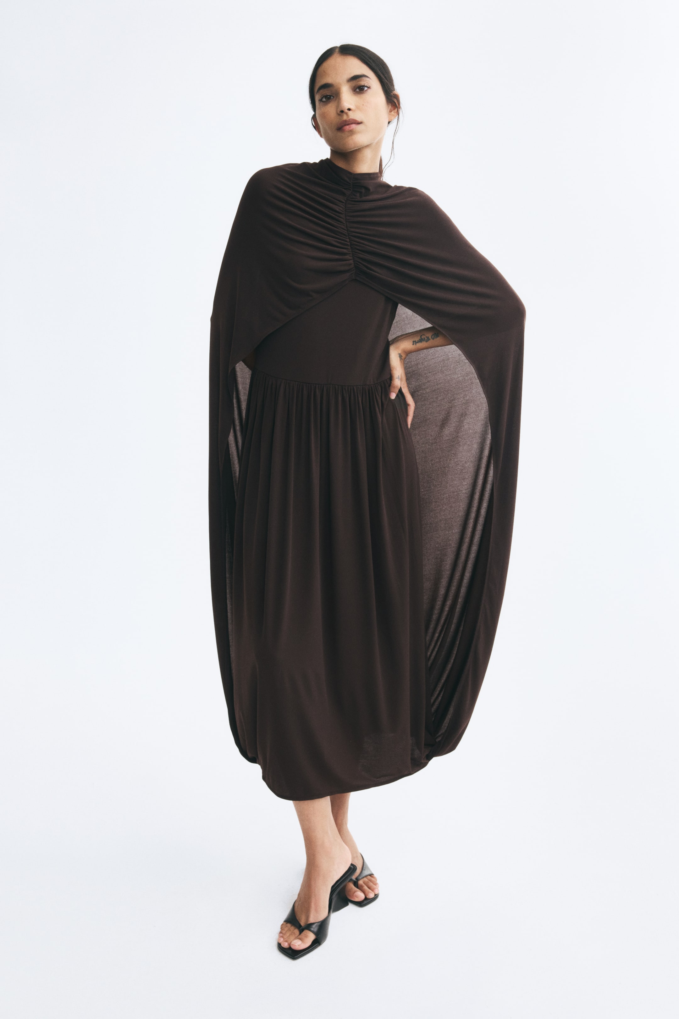 View larger image: The model is wearing a dark brown jersey dress featuring a cape-like outer layer with a gathered neckline that drapes over the shoulders and arms. The inner dress layer has a gathered waist and is styled with black heeled sandals.