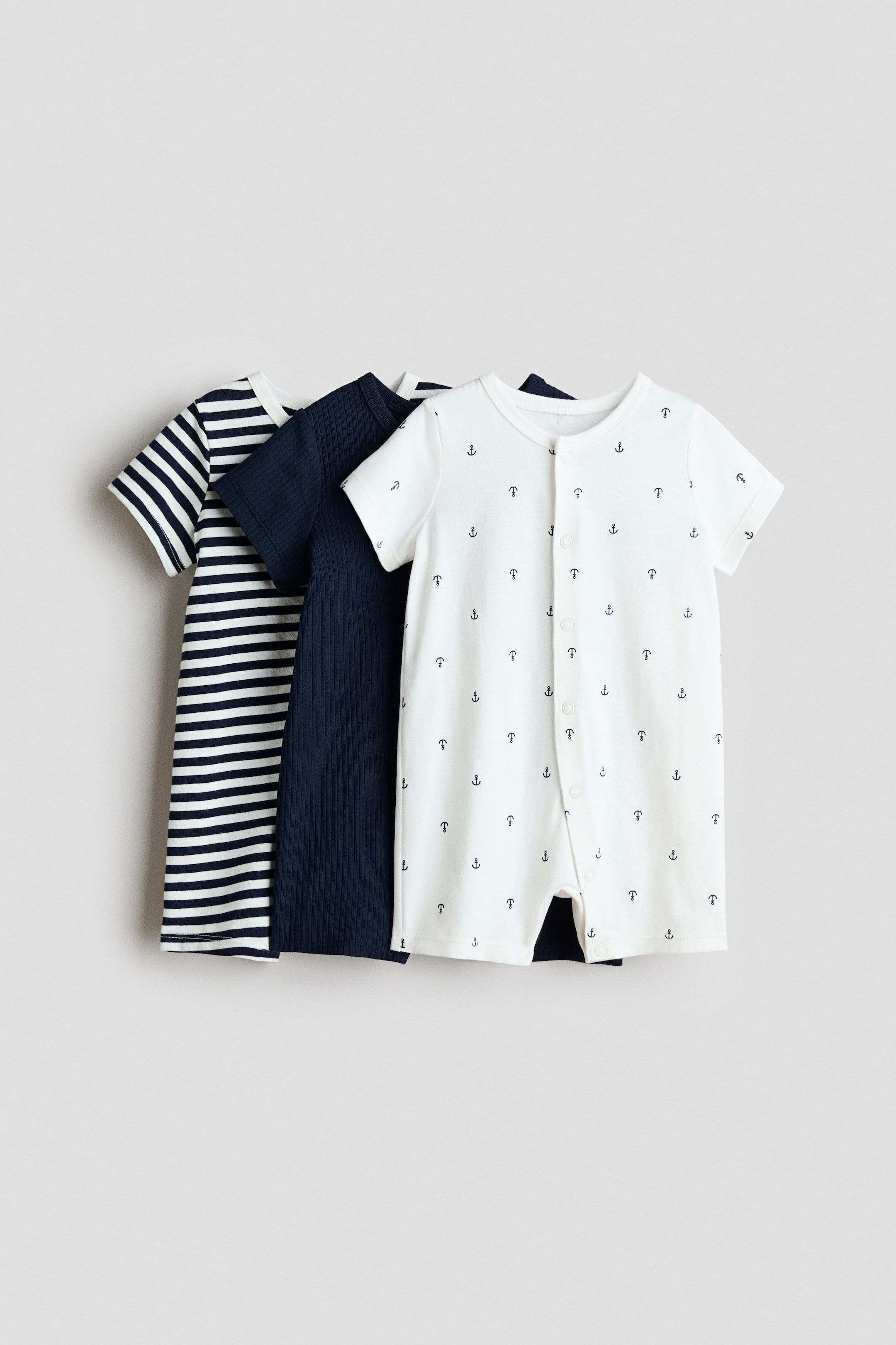 Agrandir l'image: Trois grenouillères à manches courtes en jersey de coton doux sont présentées, dotées d'un col rond, de manches courtes et de boutons-pression sur le devant. Une grenouillère est blanche avec un motif d'ancre sombre répété, une autre est bleu marine uni avec une texture côtelée, et la troisième est rayée blanc et bleu marine.