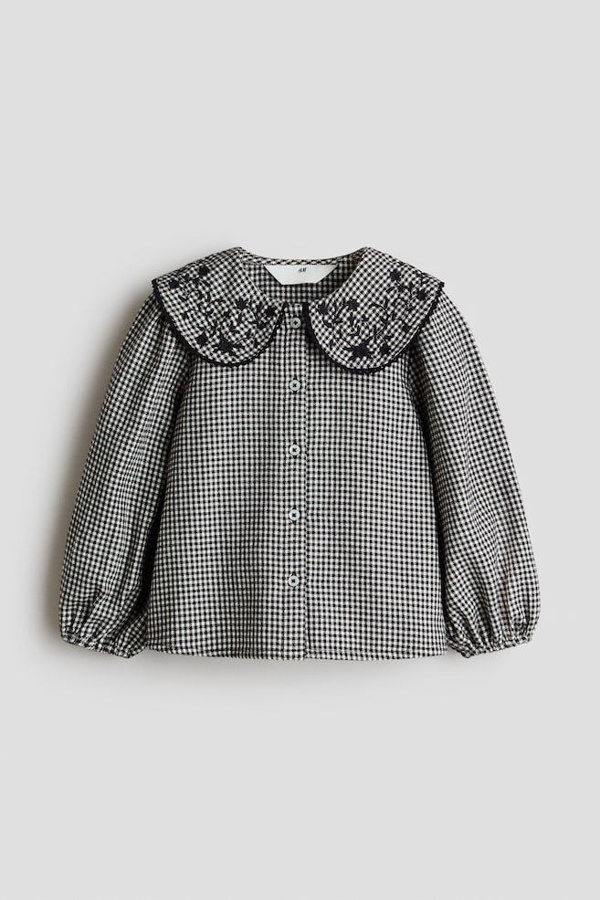 A front-facing view of a cotton twill blouse in a black-and-white houndstooth pattern, showcasing a large Peter Pan collar with black embroidered floral motifs and a picot-trimmed edge. The blouse features a button-through front and long puff sleeves with elasticated cuffs. - 1
