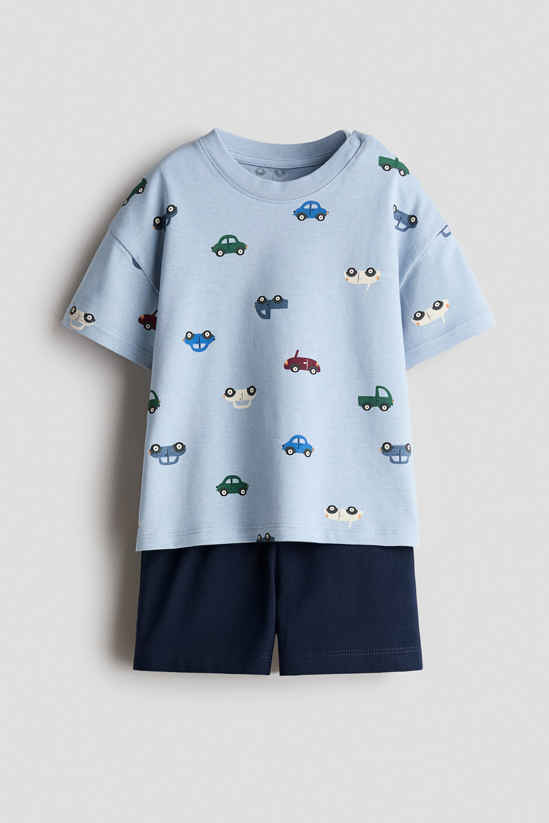 View larger image: A 2-piece cotton set is being showcased from the front. The light blue t-shirt is adorned with an allover print of vibrant cars and is crafted with short sleeves and a ribbed crew neck with snap buttons on the shoulder. The solid dark blue shorts are layered beneath the t-shirt.