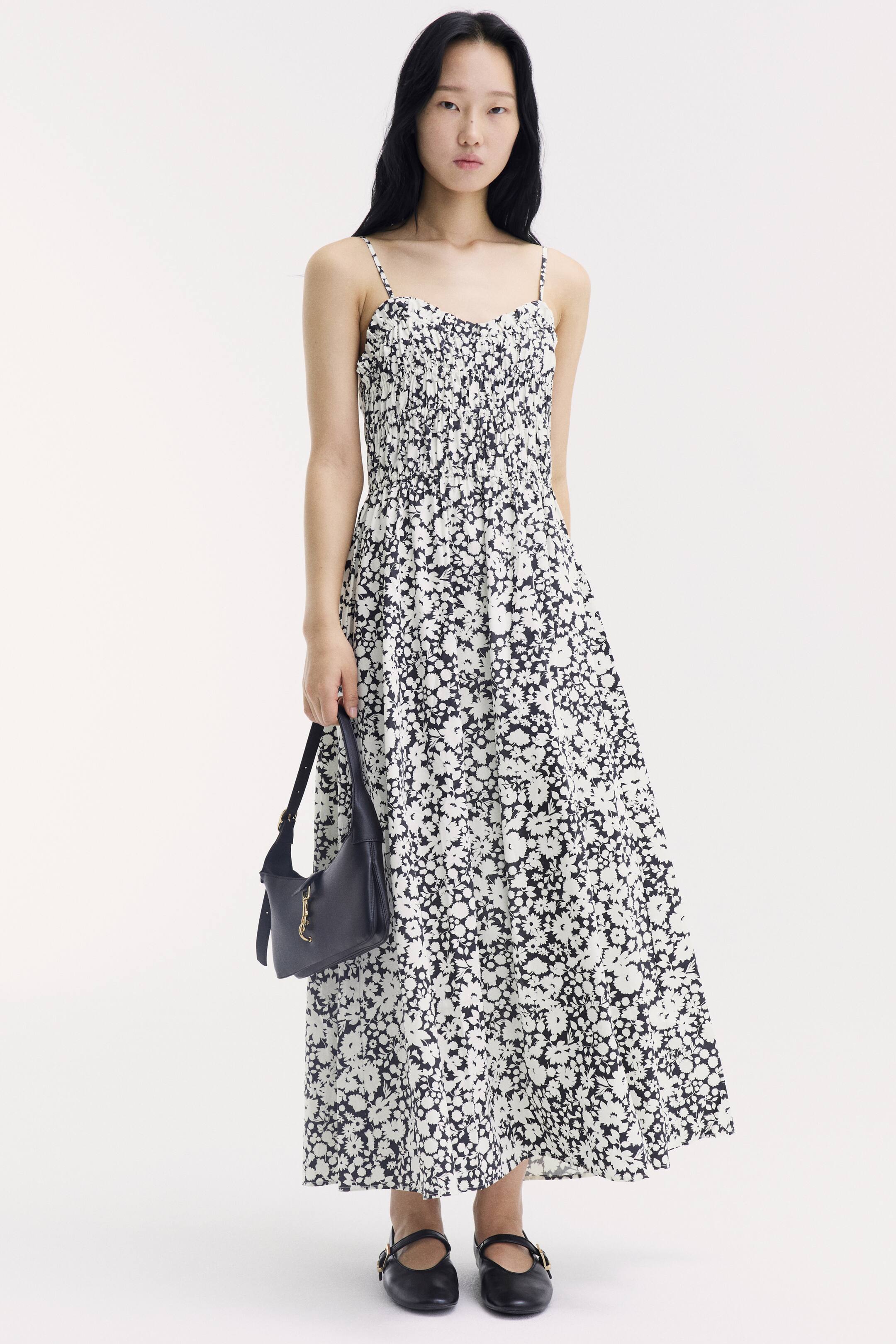 View larger image: A calf-length dress featuring a black and white floral print, thin shoulder straps, a ruched bodice, and a flared skirt is being worn by the model. The model is also holding a small black handbag and wearing black flat shoes.