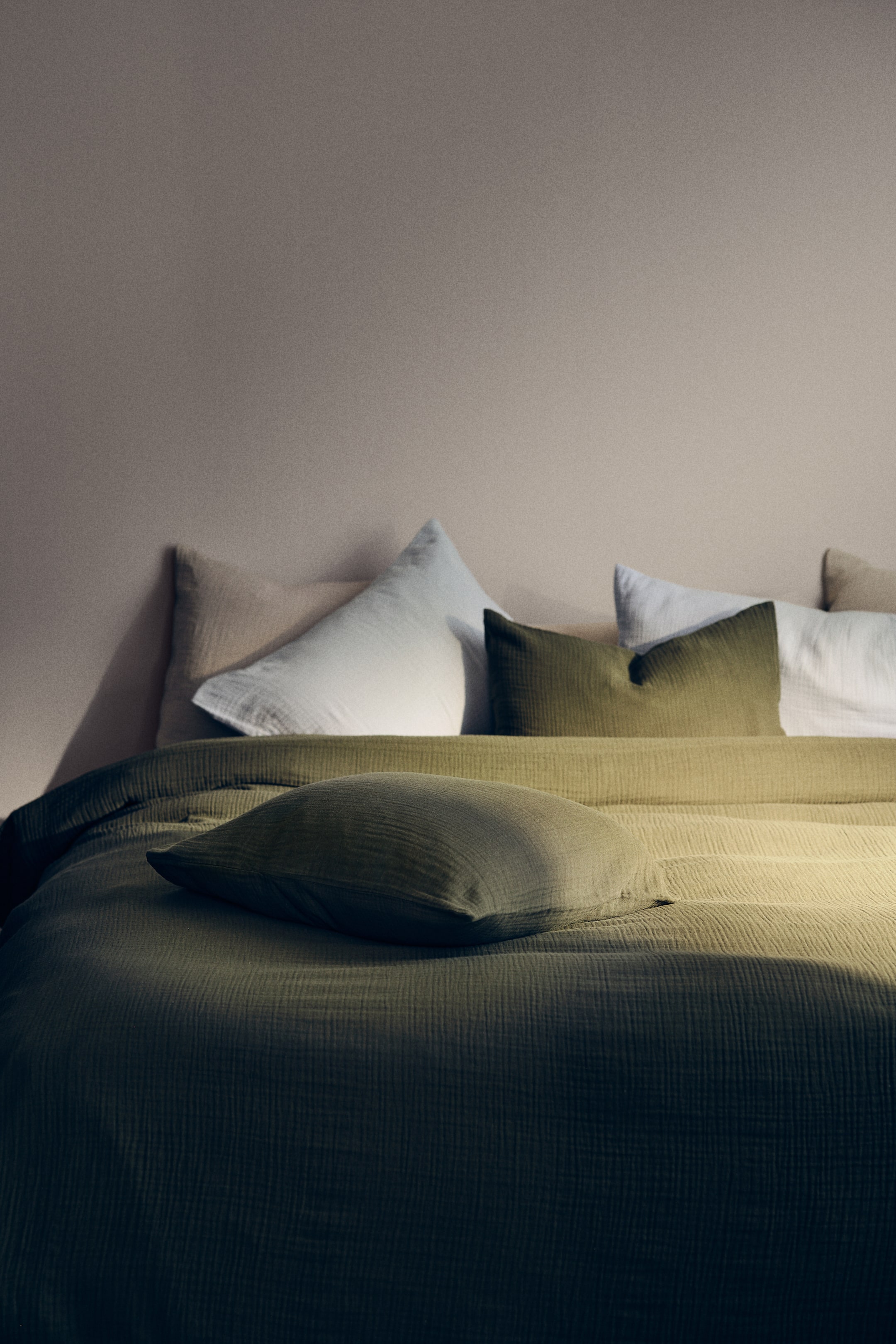 View larger image: An olive green bedding set is draped over a bed, showcasing a textured, crinkled duvet. A coordinating olive green pillow is placed on the duvet, with additional pillows in olive green, white, and light beige arranged upright at the head of the bed.
