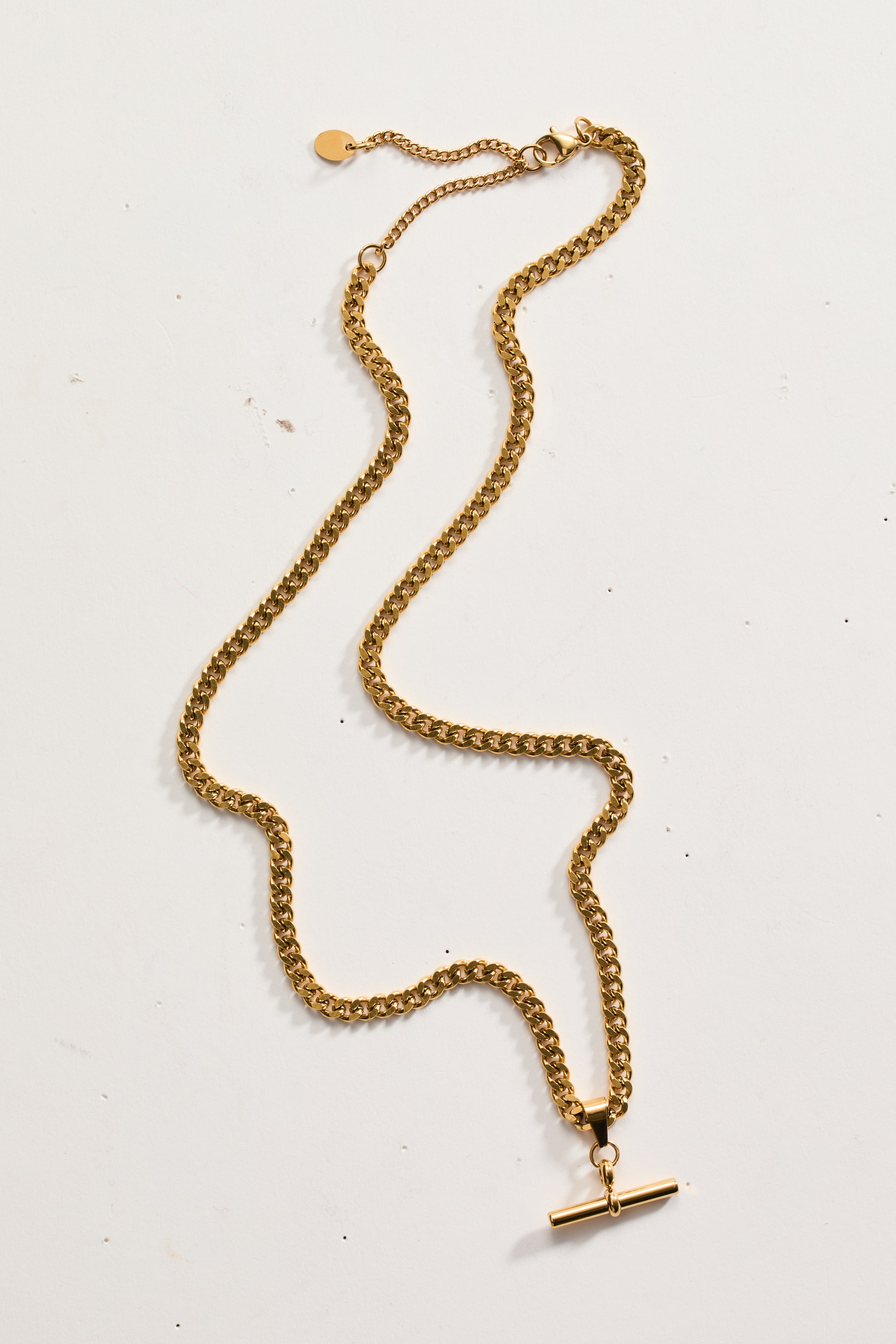 Προβολή μεγαλύτερης εικόνας: Ένα κολιέ με αλυσίδα τύπου curb σε χρυσή απόχρωση είναι τοποθετημένο επίπεδα σε σχήμα S, διαθέτοντας κούμπωμα τύπου αστακού και μια ρυθμιζόμενη αλυσίδα επέκτασης με μια μικρή στρογγυλή γοητεία στη μία άκρη. Ένα κούμπωμα τύπου T-bar toggle είναι προσαρτημένο στην κύρια αλυσίδα στην άλλη άκρη.