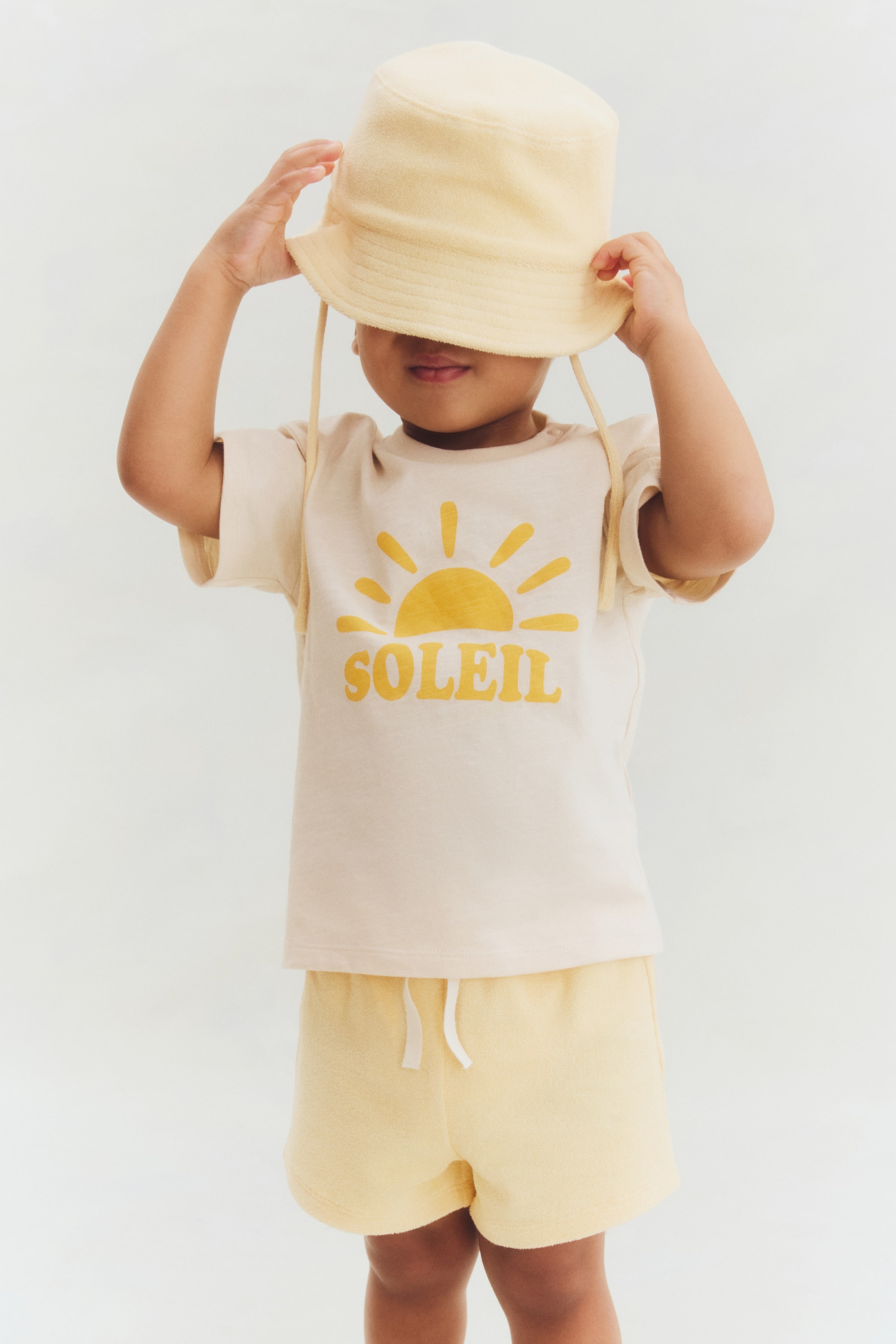 Grotere afbeelding bekijken: Een lichtgele badstof vissershoed met kinbanden wordt gedragen, gecombineerd met een crèmekleurig t-shirt met korte mouwen met een gele 'SOLEIL' zon graphic en bijpassende lichtgele badstof shorts met een witte trekkoord.