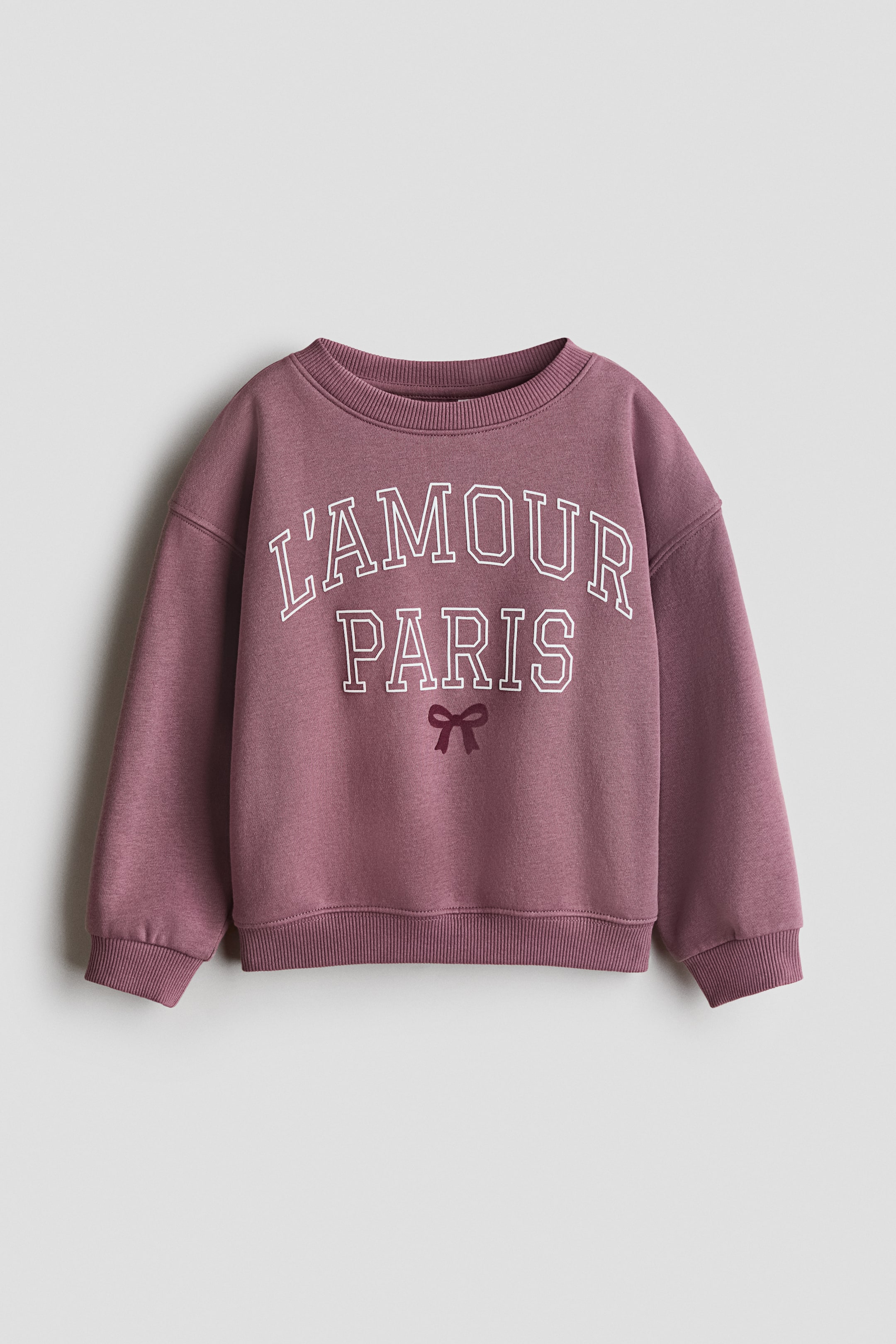 View larger image: A muted purple top in printed sweatshirt fabric is lying flat, facing forward. It features a round, rib-trimmed neckline and dropped shoulders. The front displays "L'AMOUR PARIS" in white outlined letters with a small dark plum bow graphic below, and ribbed cuffs and a ribbed hem complete the design.