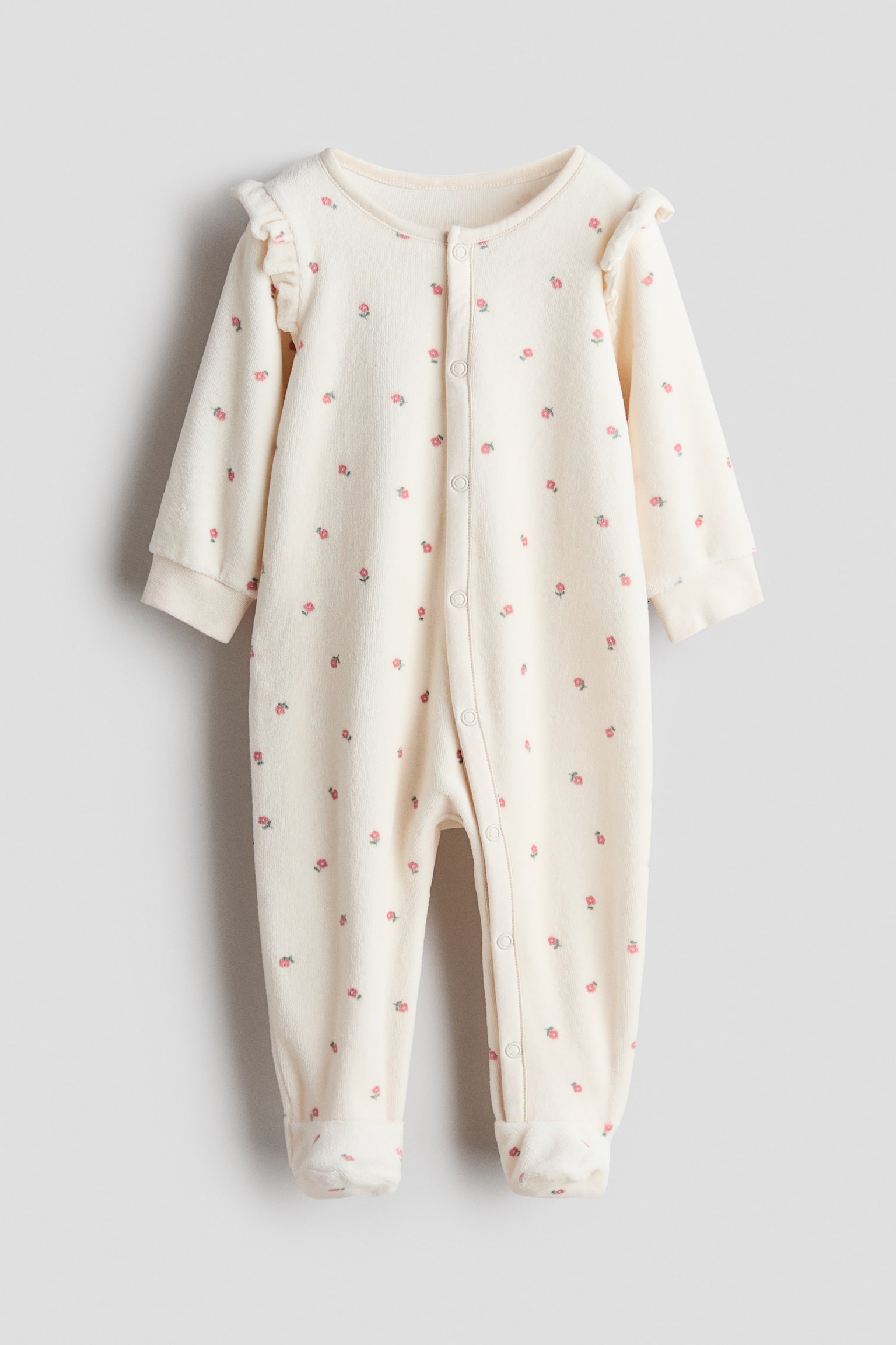 A front view of a velour sleepsuit featuring an off-white fabric with a small, repeating pink floral pattern. The design features ruffle details at the shoulders, a snap-button closure extending down the centre front and one leg, ribbed cuffs, and fully enclosed feet.