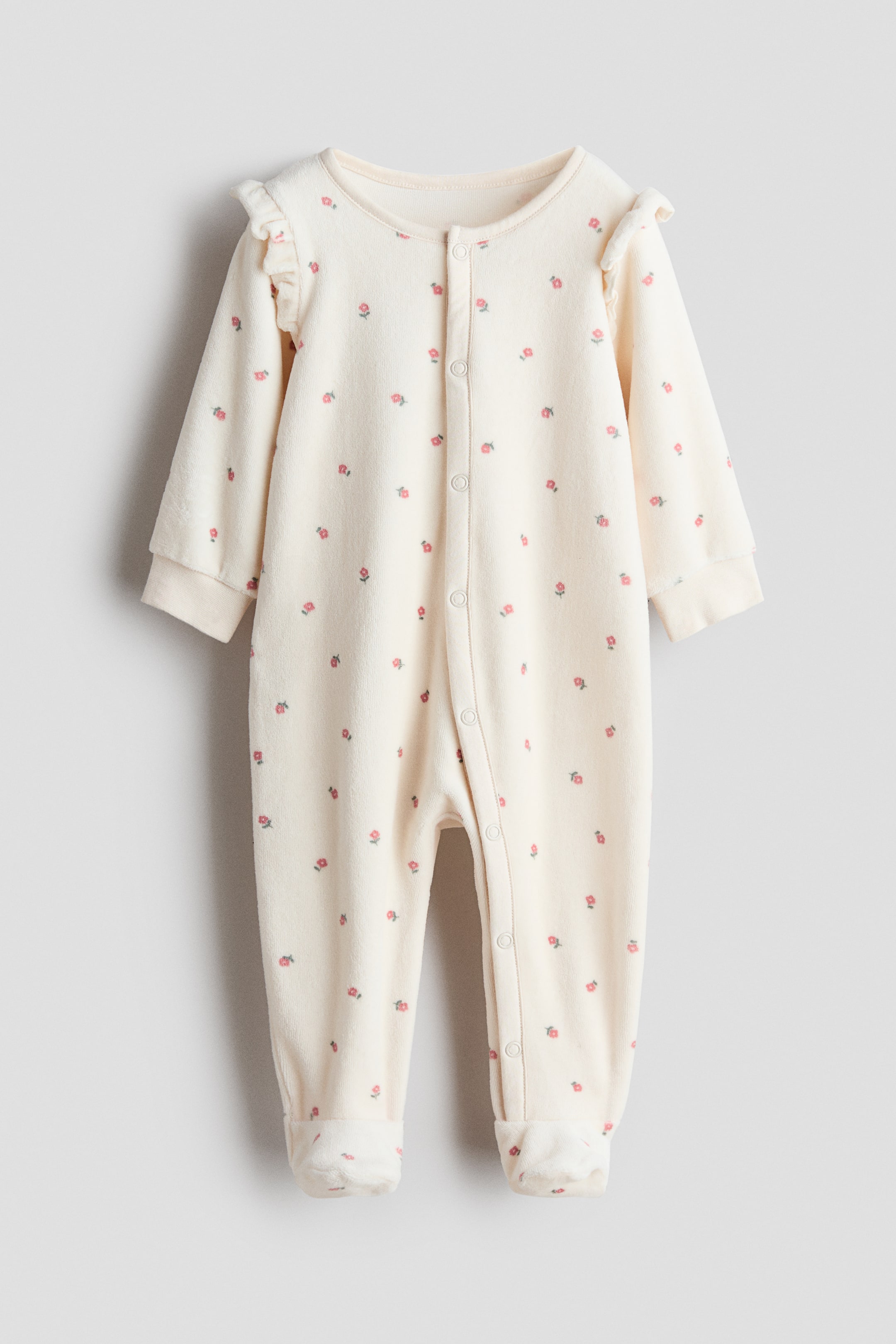 View larger image: A front view of a velour sleepsuit showing off-white fabric with a small, repeating pink floral pattern. The design features ruffle details at the shoulders, a snap-button closure extending down the center front and one leg, ribbed cuffs, and fully enclosed feet.