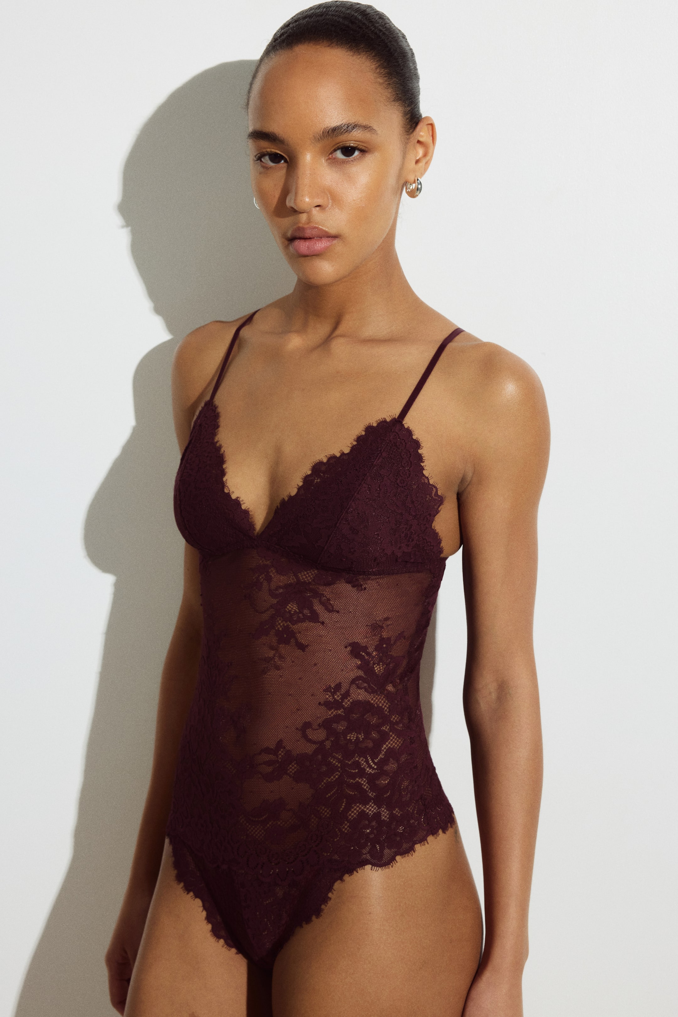 View larger image: The model is wearing a dark red V-neck thong bodysuit in lace with adjustable shoulder straps and lined cups. The bodysuit features sheer lace with a floral pattern.