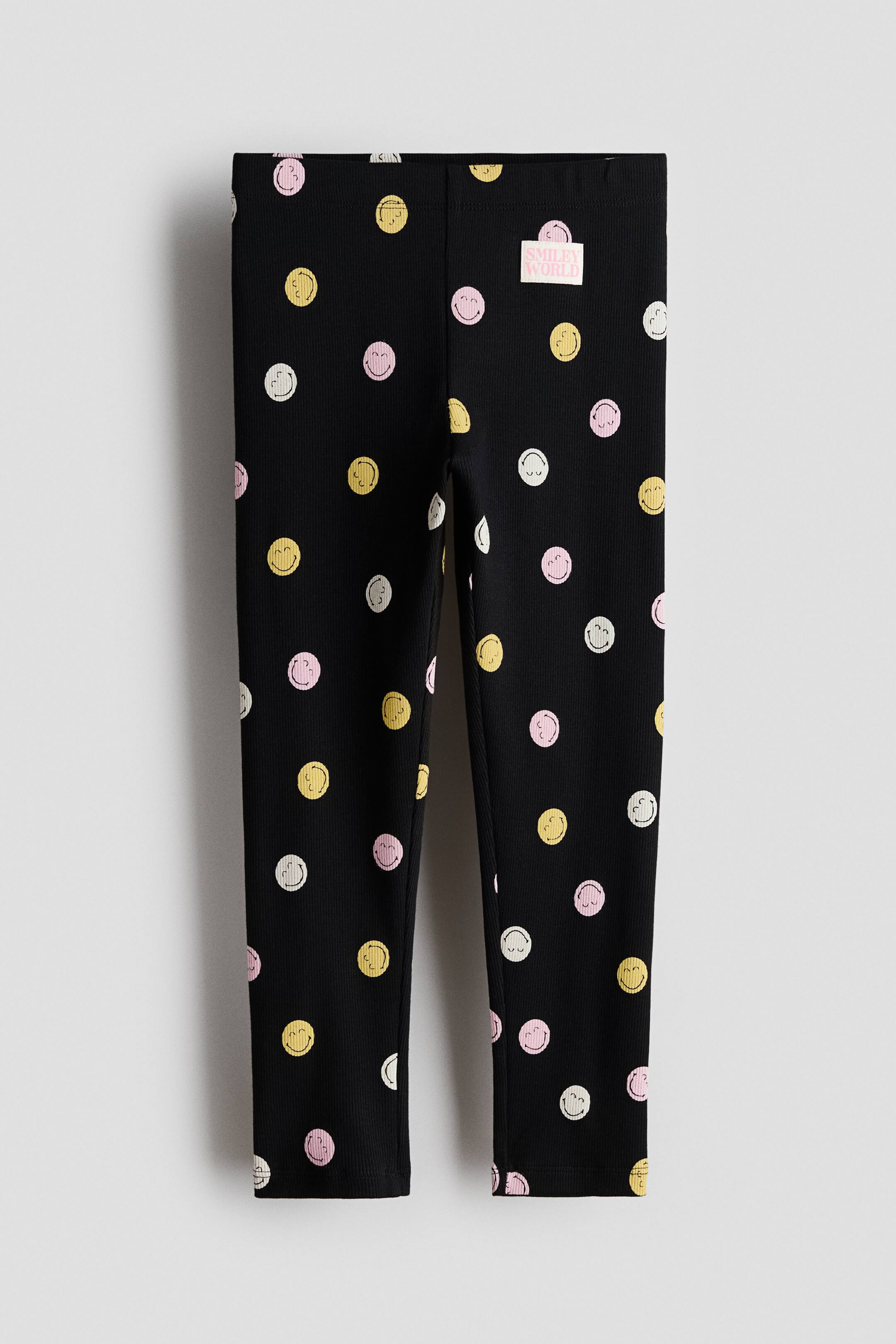 Büyük resmi görüntüle: Siyah nervürlü taytların önden görünümü gösteriliyor; tüm yüzeyi sarı, pembe ve beyaz smiley yüzleriyle kaplı bir desen içeriyor. Bel bandının sağ tarafında beyaz "SMILEY WORLD" etiketi görülüyor.