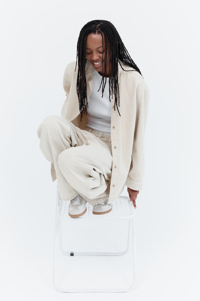 An oversized, light beige denim shirt is worn open by the model, who is crouching on a white metal stool. The shirt features a collar, front button closures, dropped shoulders, and long sleeves, styled over a white T-shirt and matching light-coloured jeans. - 1