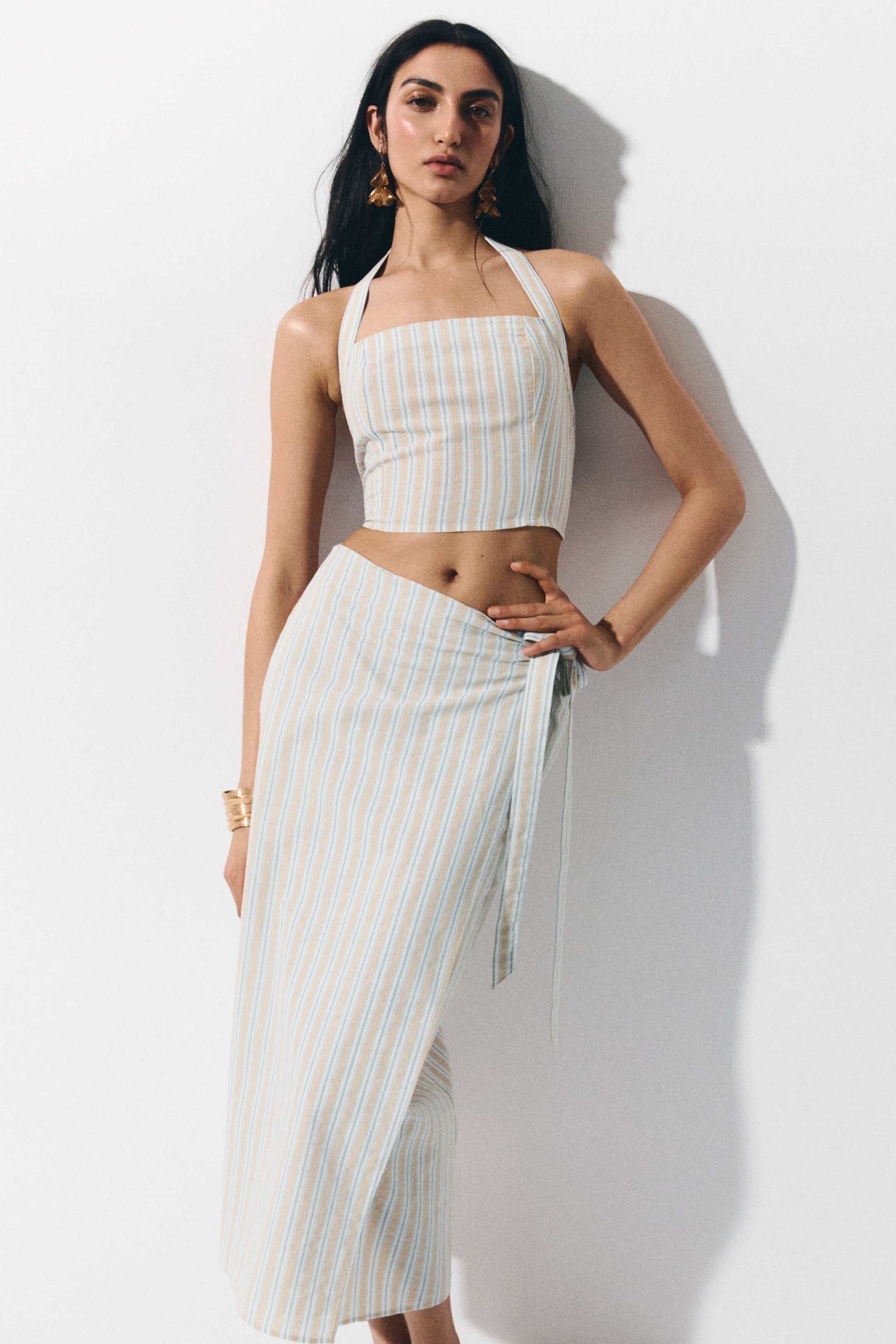 Ampliar la imagen: El producto es un conjunto de dos piezas, que consta de un top corto con cuello halter y una falda envolvente a juego, ambos con rayas verticales en tonos bronceado y azul claro. La falda envolvente se anuda al costado, formando un frente asimétrico.