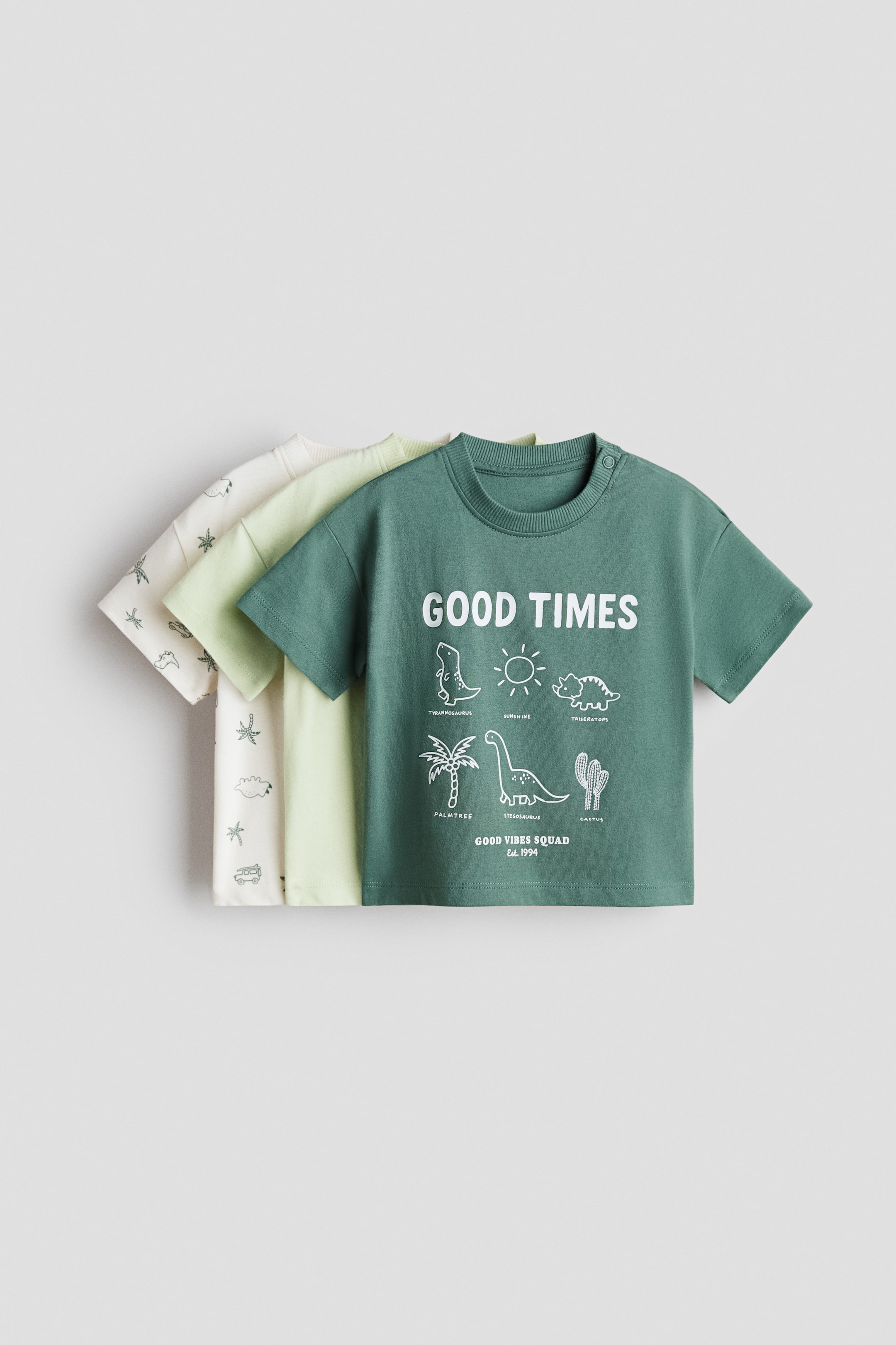 View larger image: A 3-pack of cotton T-shirts is presented, featuring three short-sleeved shirts layered. The frontmost dark green T-shirt faces forward, showing a ribbed crew neck with two snap buttons on the left shoulder and a white graphic print reading 'GOOD TIMES' with illustrations of dinosaurs, the sun, a palm tree and a cactus. Behind it, a light green T-shirt is partially visible, followed by an off-white T-shirt at the back with a subtle pattern of stars and clouds.
