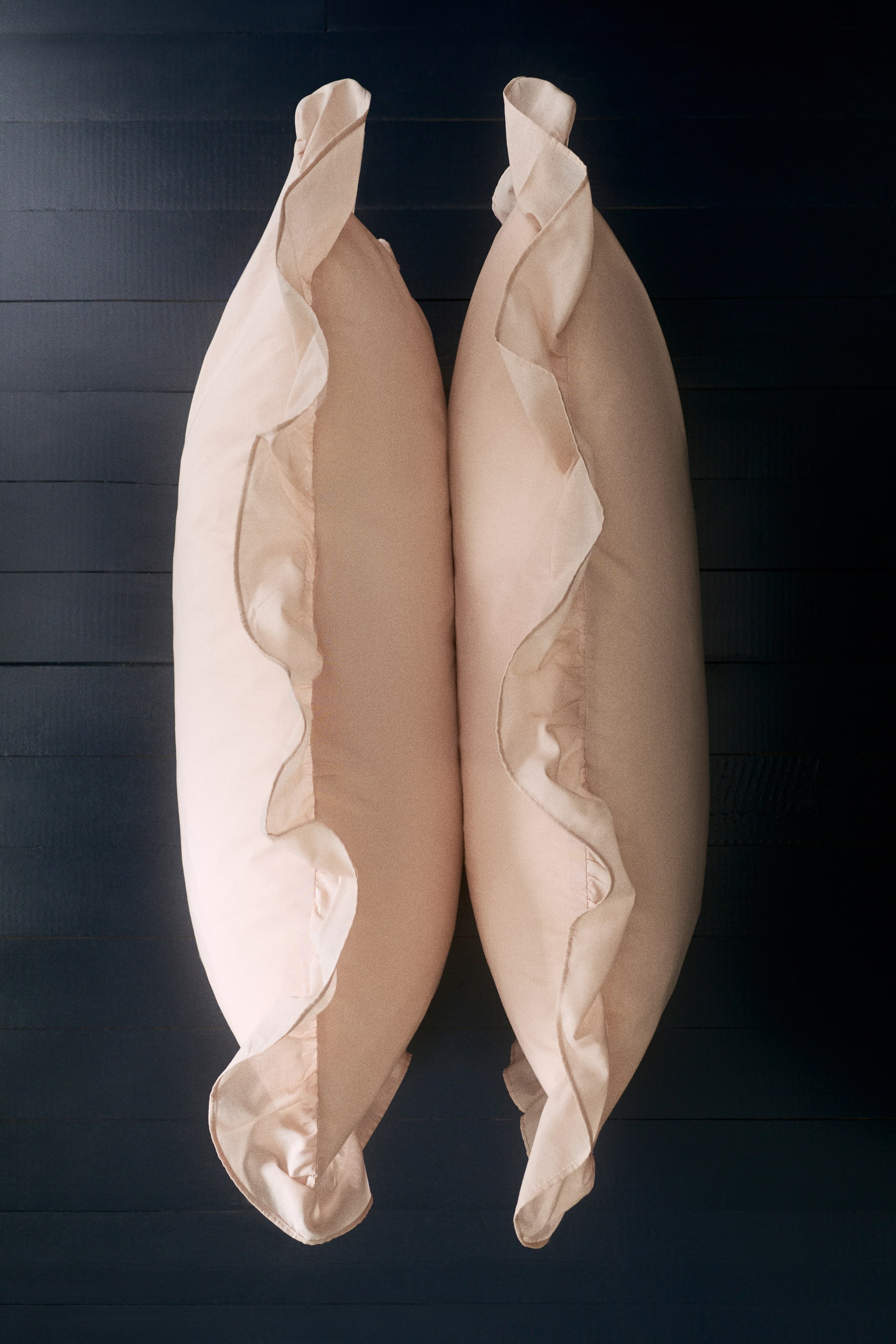 이미지 확대 보기: 두 개의 연한 복숭아색 베개 커버가 나란히 놓여 있으며, 각각의 가시적인 가장자리를 따라 넓은 주름 장식이 특징입니다.