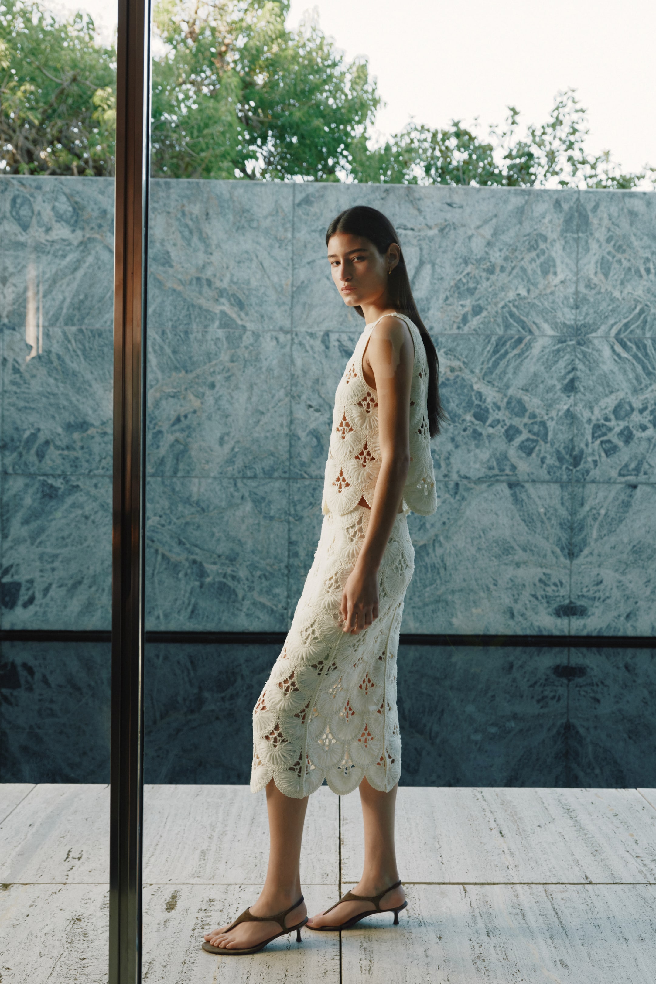 View larger image: A cream crocheted sleeveless top paired with a matching midi skirt is showcased in a side profile, highlighting the repeating scalloped openwork patterns throughout. The sleeveless top finishes above the waist, while the midi skirt features a scalloped hemline, extending below the knee. Dark brown kitten heel thong sandals complete the ensemble.