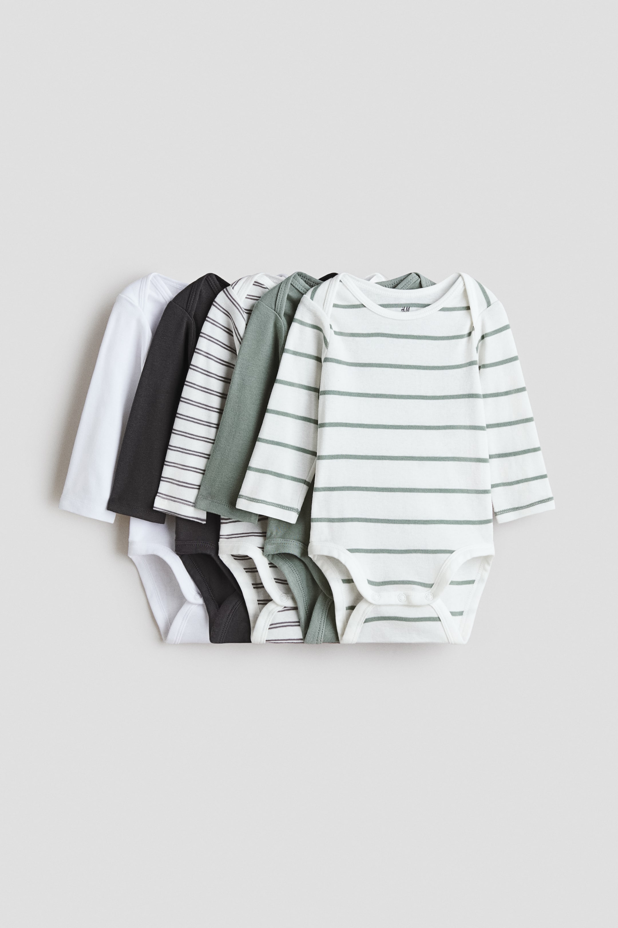 Grotere afbeelding bekijken: Vijf babypakjes van katoenjersey met lange mouwen liggen overlappend in een frontale weergave, met envelophalslijnen en drukknoopsluitingen aan de onderkant. De collectie bestaat uit een mix van effen en gestreepte designs in wit, donkergrijs en saliegroen.