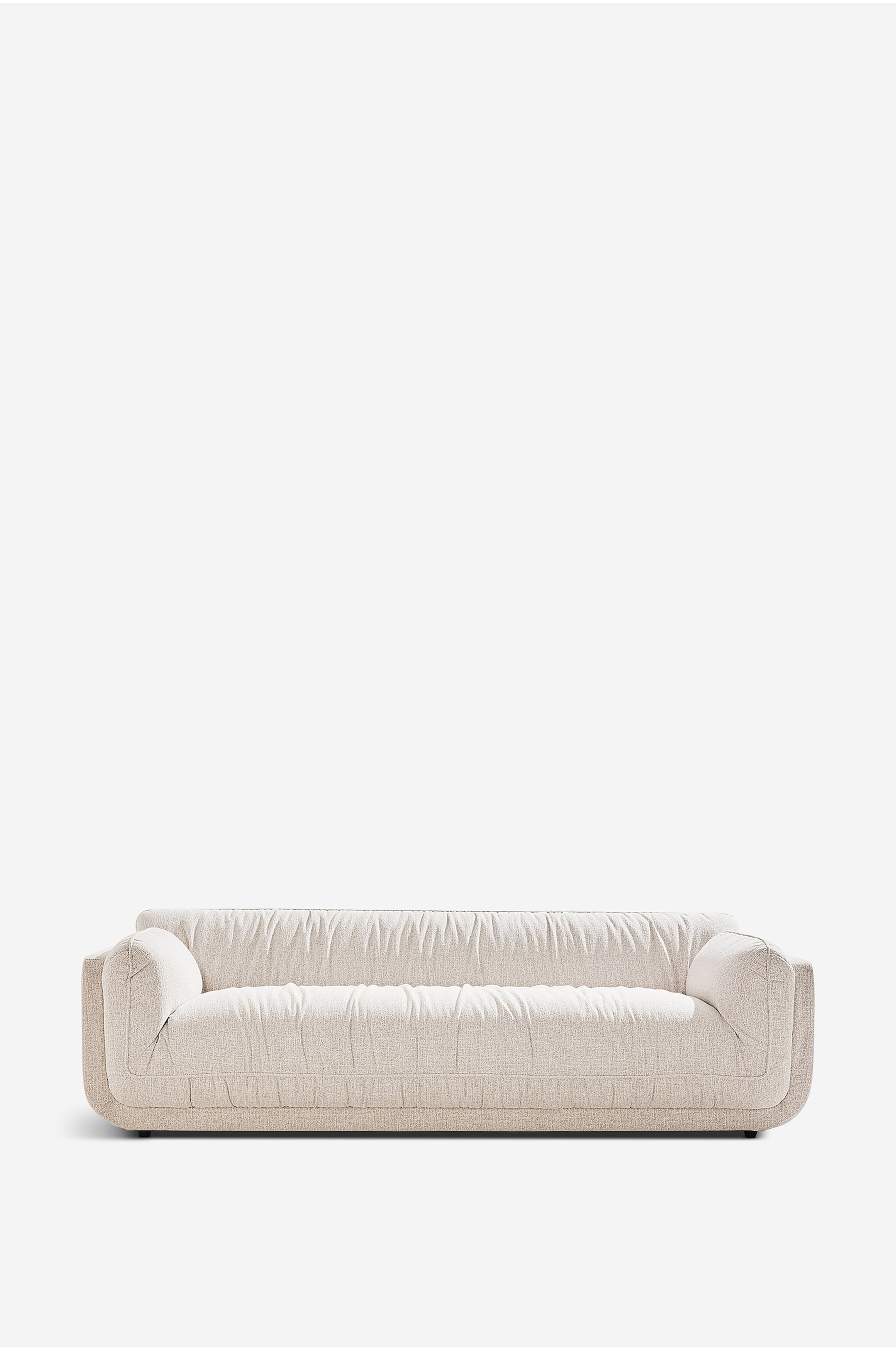 Visa större bild: En ljusbeige soffa står framåt och visar upp en mjuk, voluminös form med breda, rundade armstöd samt ett ryggstöd och en sits täckta i rynkad, texturerad tyg.