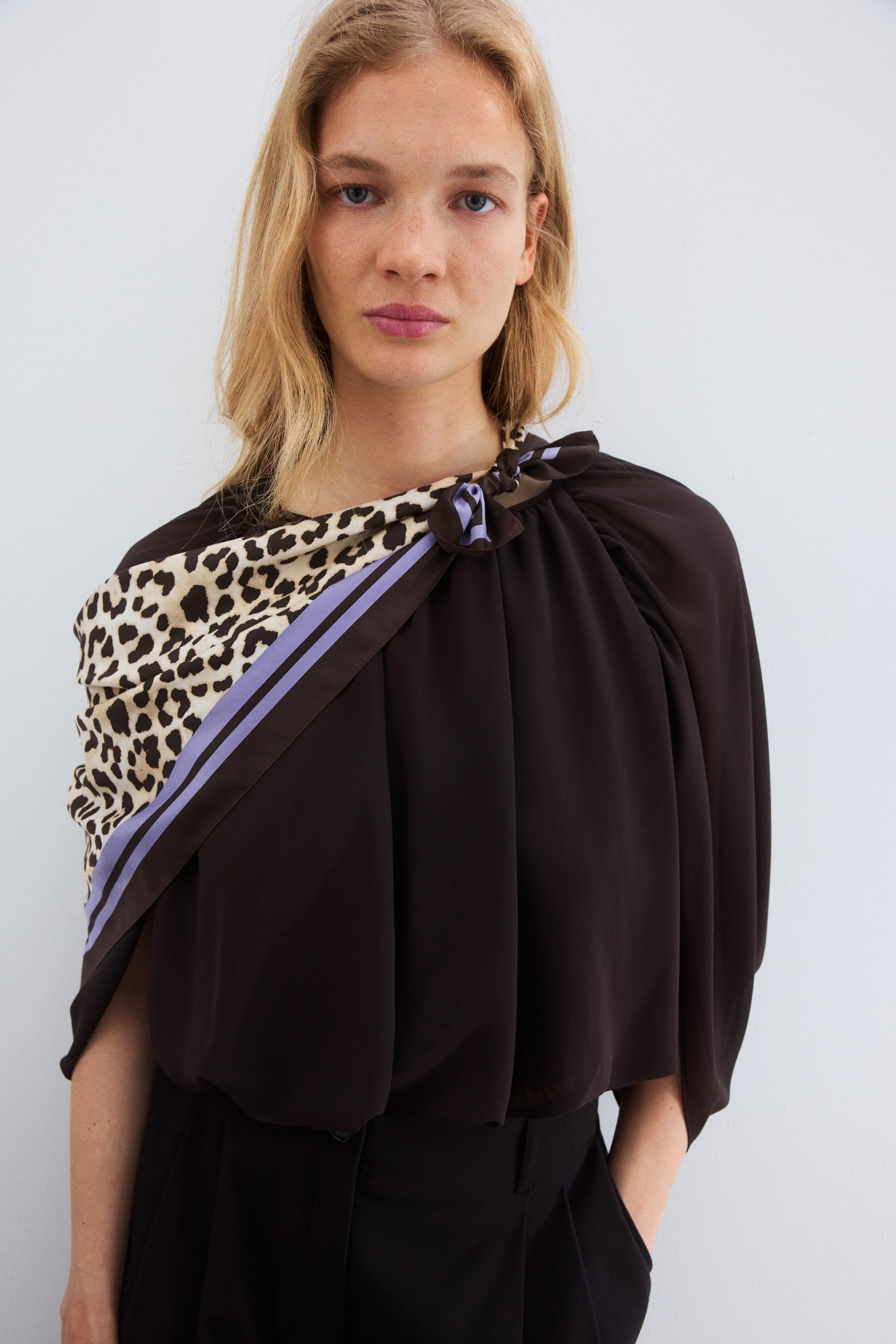 Vis større bilde: En mørkebrun bluse med drapert silhuett, med et skjerf i leopardmønster som knytes på høyre skulder. Skjerfet har parallelle lavendelfargede striper og en brun kant, som skaper en asymmetrisk utringning.