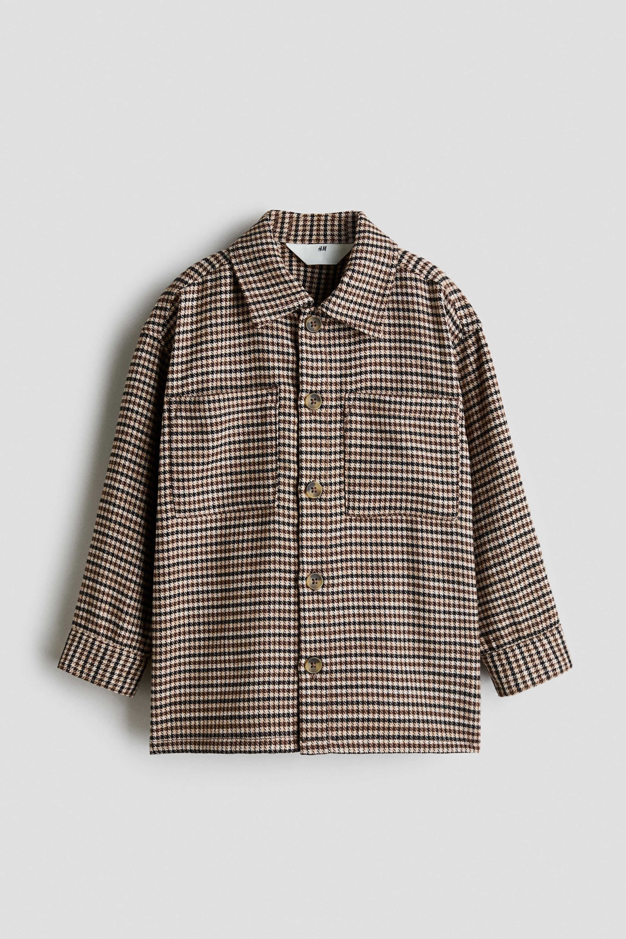 View larger image: An oversized overshirt in a brown, black, and beige houndstooth pattern is shown from the front, featuring a turn-down collar, a button-front closure, and two patch chest pockets. The design includes dropped shoulders and long sleeves with wide cuffs.