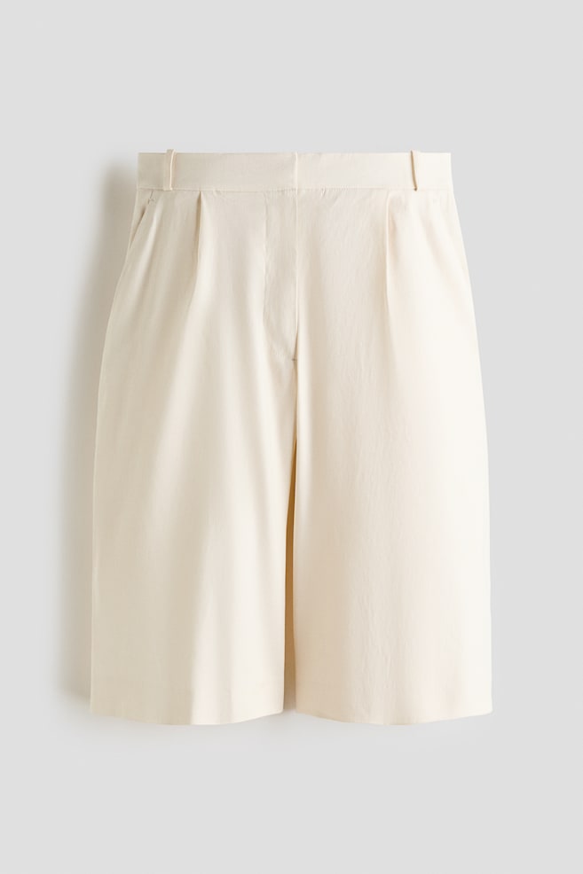 Les shorts crème sont présentés de face, avec une ceinture plate dotée de passants et des plis marqués à l'avant, s'élargissant en une coupe large qui se termine au-dessus du genou. - 1