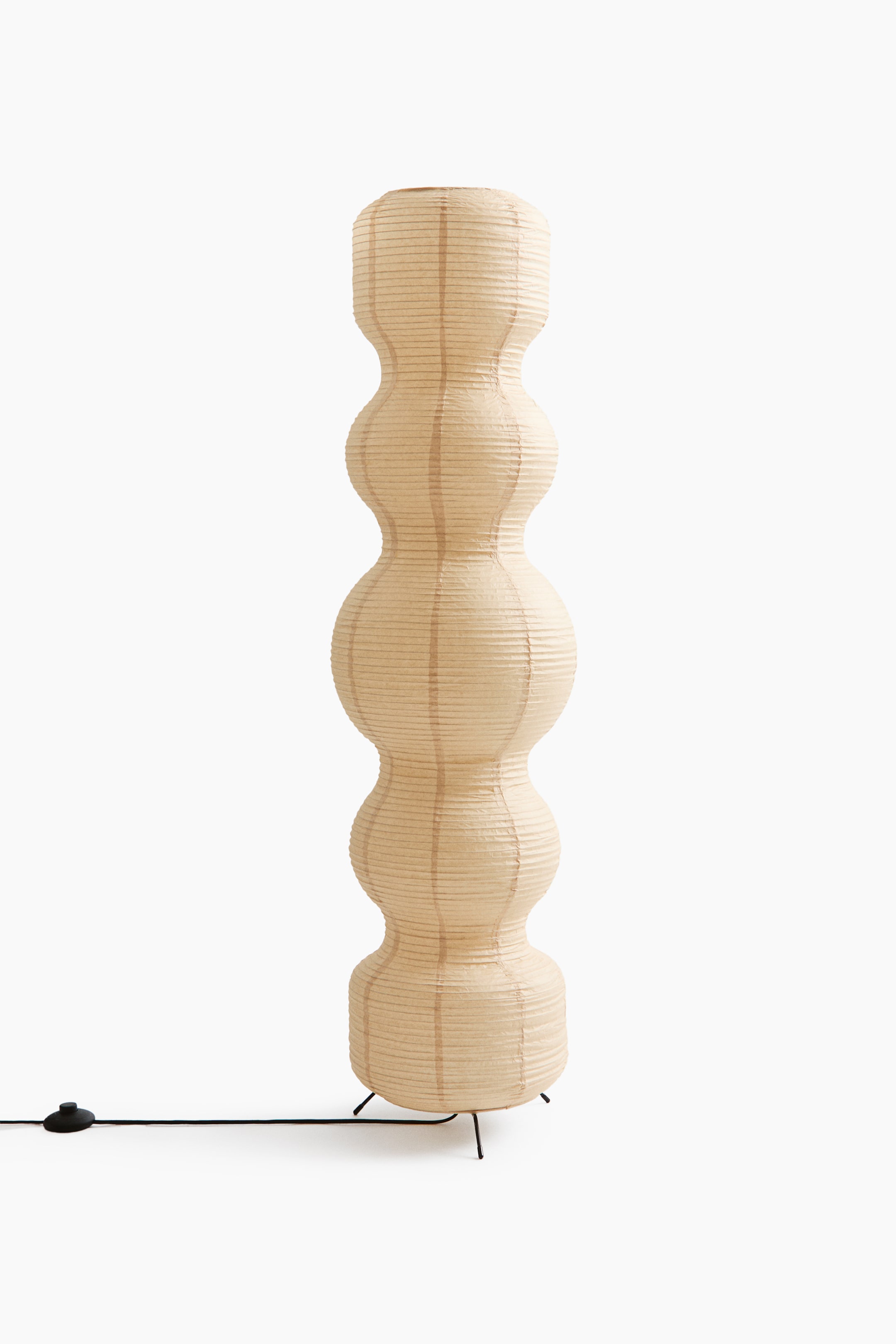 View larger image: A floor lamp stands on slim black metal legs, featuring a tall, rippling rice paper shade with five bulbous sections in a cream hue. A black fabric-covered power cord extends from the lamp base and includes a round foot switch.