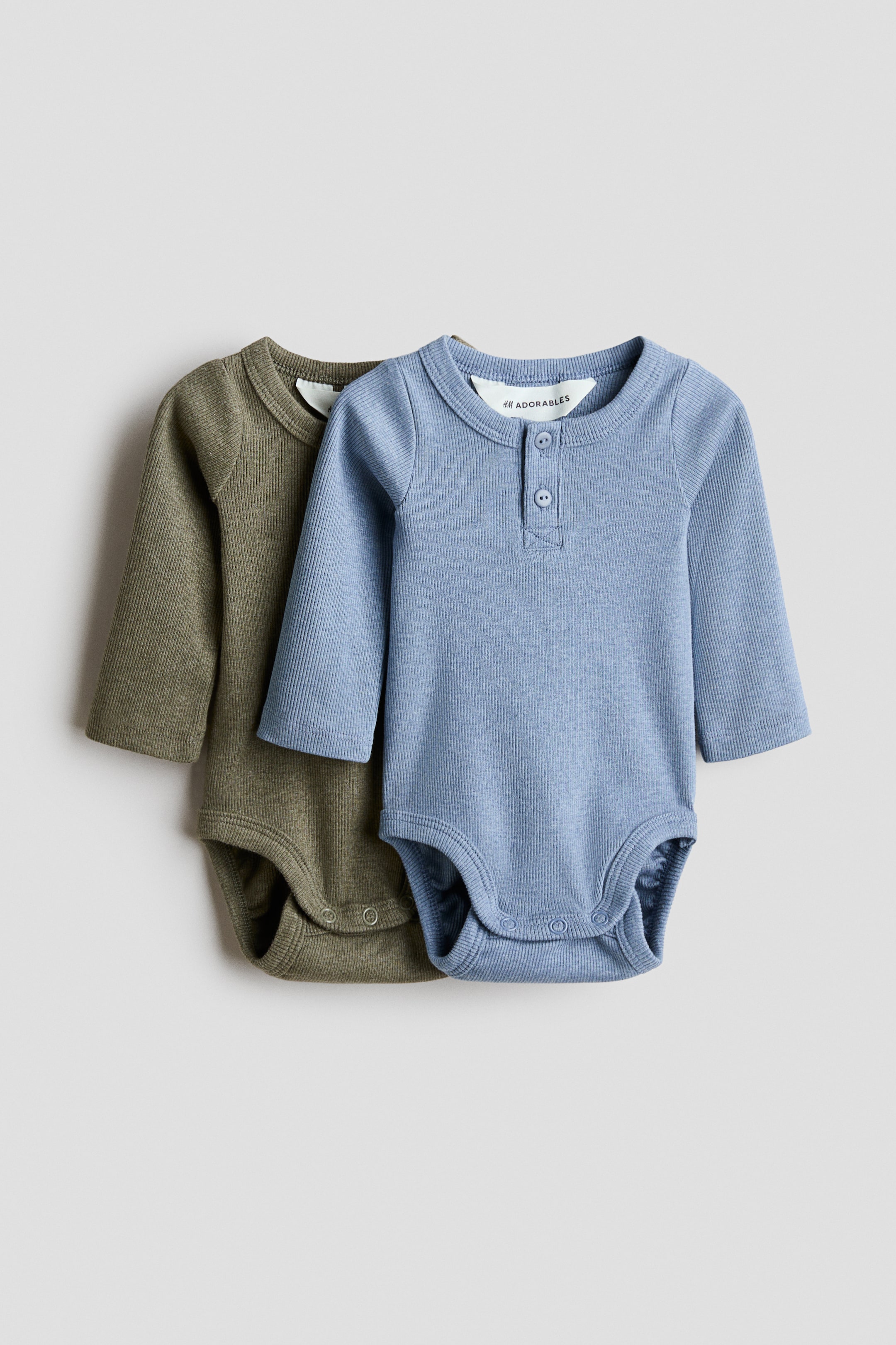 View larger image: Two baby bodysuits feature a ribbed knit texture and long sleeves. The light blue bodysuit at the front has a round neckline with a two-button Henley placket and popper fastenings at the crotch, while the olive green bodysuit behind it features a round neckline, long sleeves, and popper fastenings.