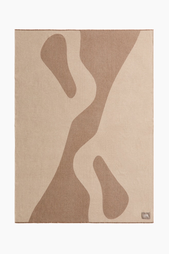 Une couverture présente un motif abstrait bicolore de formes ondulées organiques en beige clair et marron, présentée à plat. Elle est agrémentée de bords à franges et comprend une petite étiquette de marque dans le coin inférieur droit. - 1