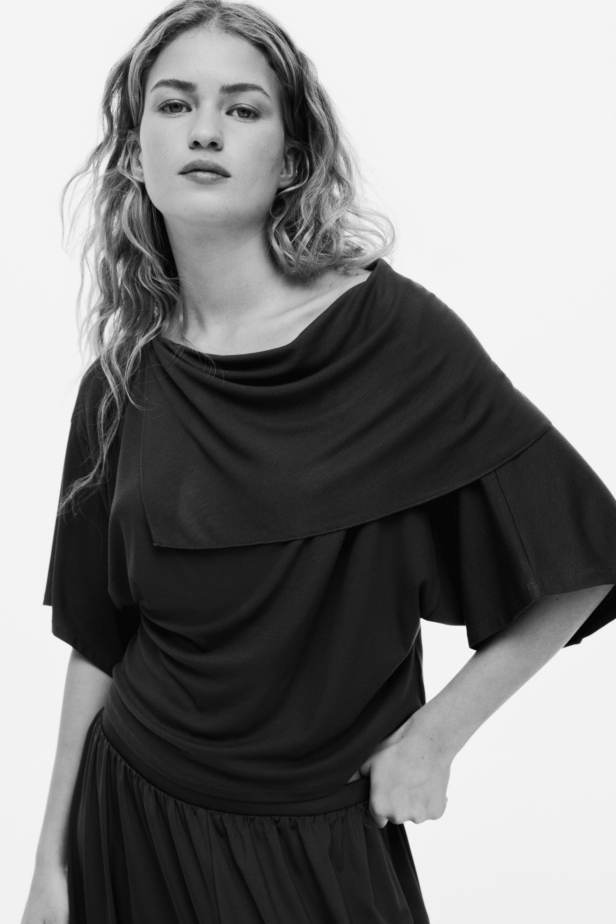 View larger image: A dark, short-sleeved top is showcased in a front-left view, featuring a wide, asymmetric cowl neckline and a relaxed fit. The top displays a layered panel across the chest and shoulders, extending into wide sleeves, and rests at the waistline of a matching dark bottom garment with a gathered elastic waistband.