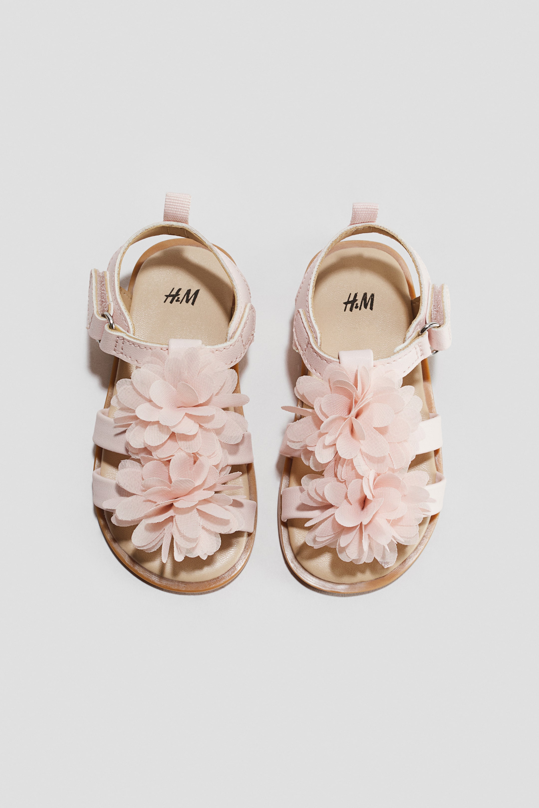 View larger image: A pair of pink children's sandals featuring multiple straps across the foot and an adjustable ankle strap with a buckle closure. Large, layered fabric floral appliqués adorn the front straps, and the H&M logo is visible on the light-coloured insole of each sandal.