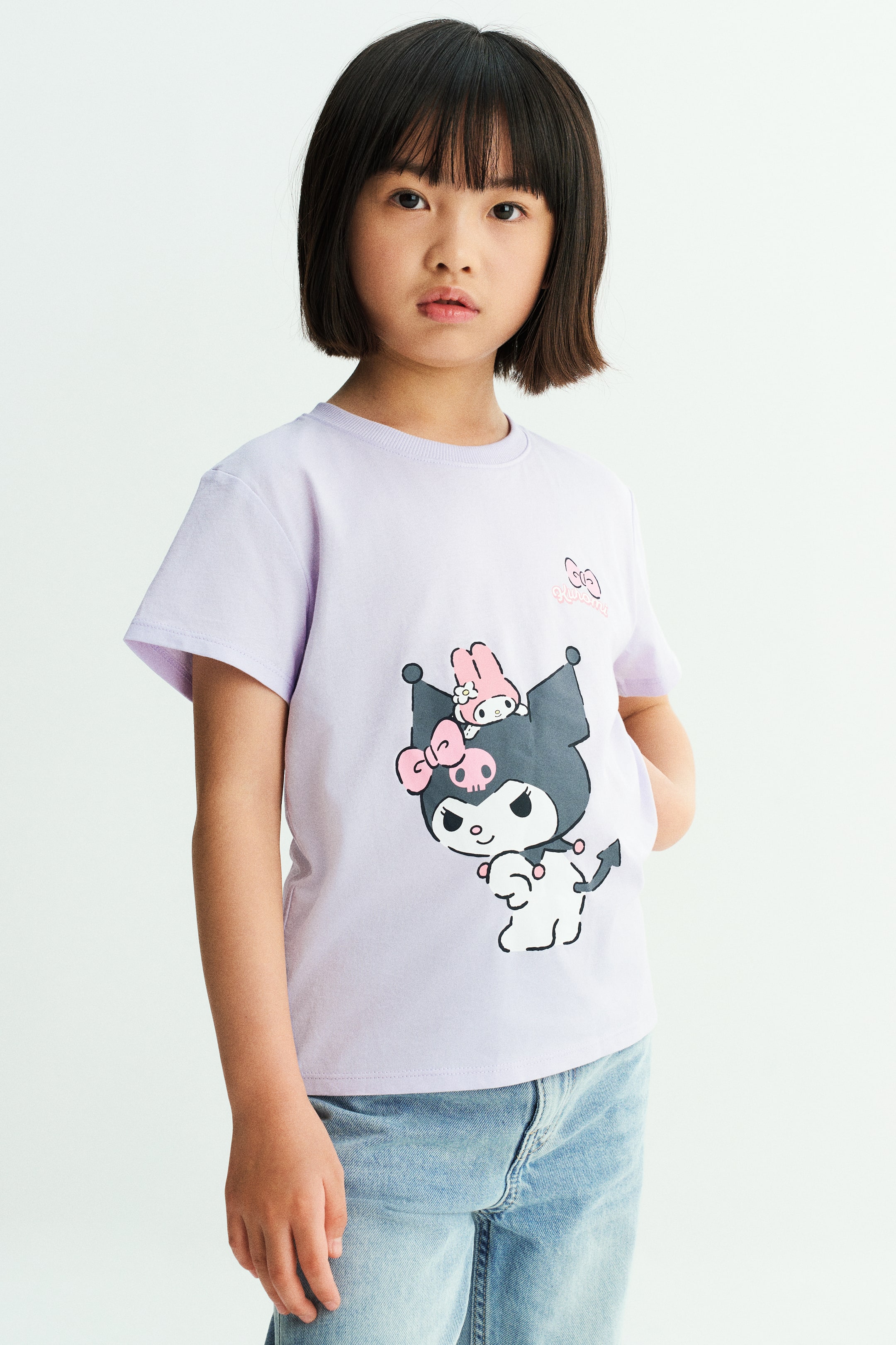 View larger image: A light purple cotton T-shirt featuring a ribbed round neckline, dropped shoulders, and a graphic character print at the front is worn by the model. The T-shirt is styled with light-wash denim jeans.