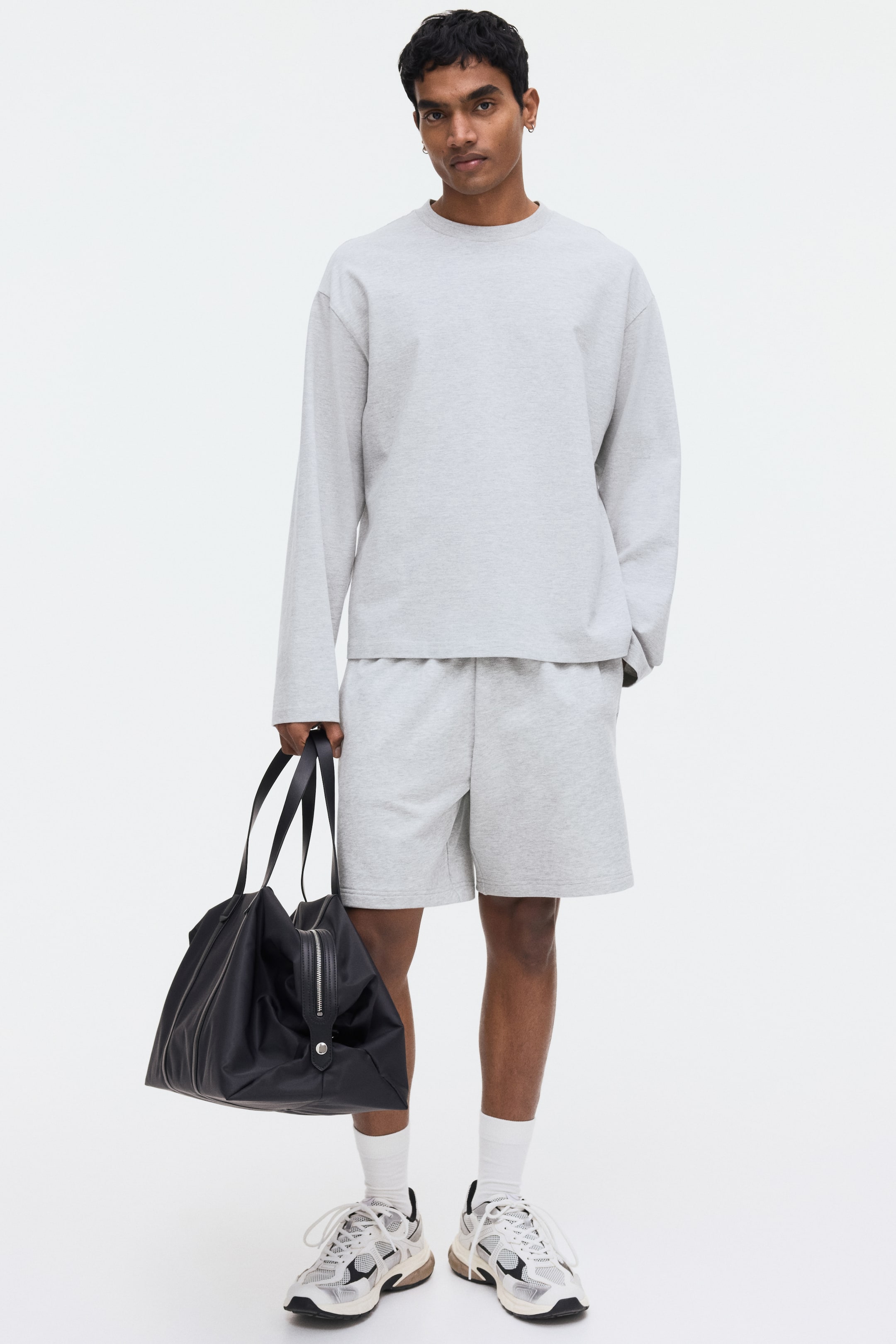 View larger image: The model is wearing light gray shorts crafted from lightweight sweatshirt fabric. These shorts have an elastic waistband and side pockets, with the model's right hand casually placed in one.