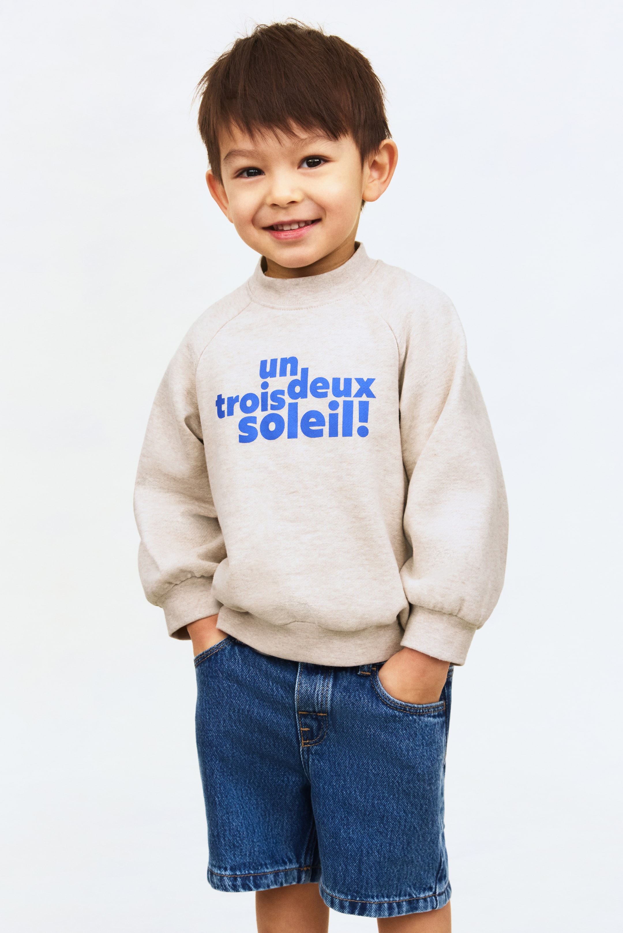 이미지 확대 보기: 정면을 향해 놓인 밝은 베이지 헤더 처리된 스웨트셔츠는 모크넥과 긴 래글런 소매, 시보리 처리된 소맷단이 특징이며 앞면에 파란색 문구가 표시되어 있습니다. 첫 번째 줄에는 "un,"이 있고 두 번째 줄에는 "trois deux soleil!"이 있습니다.