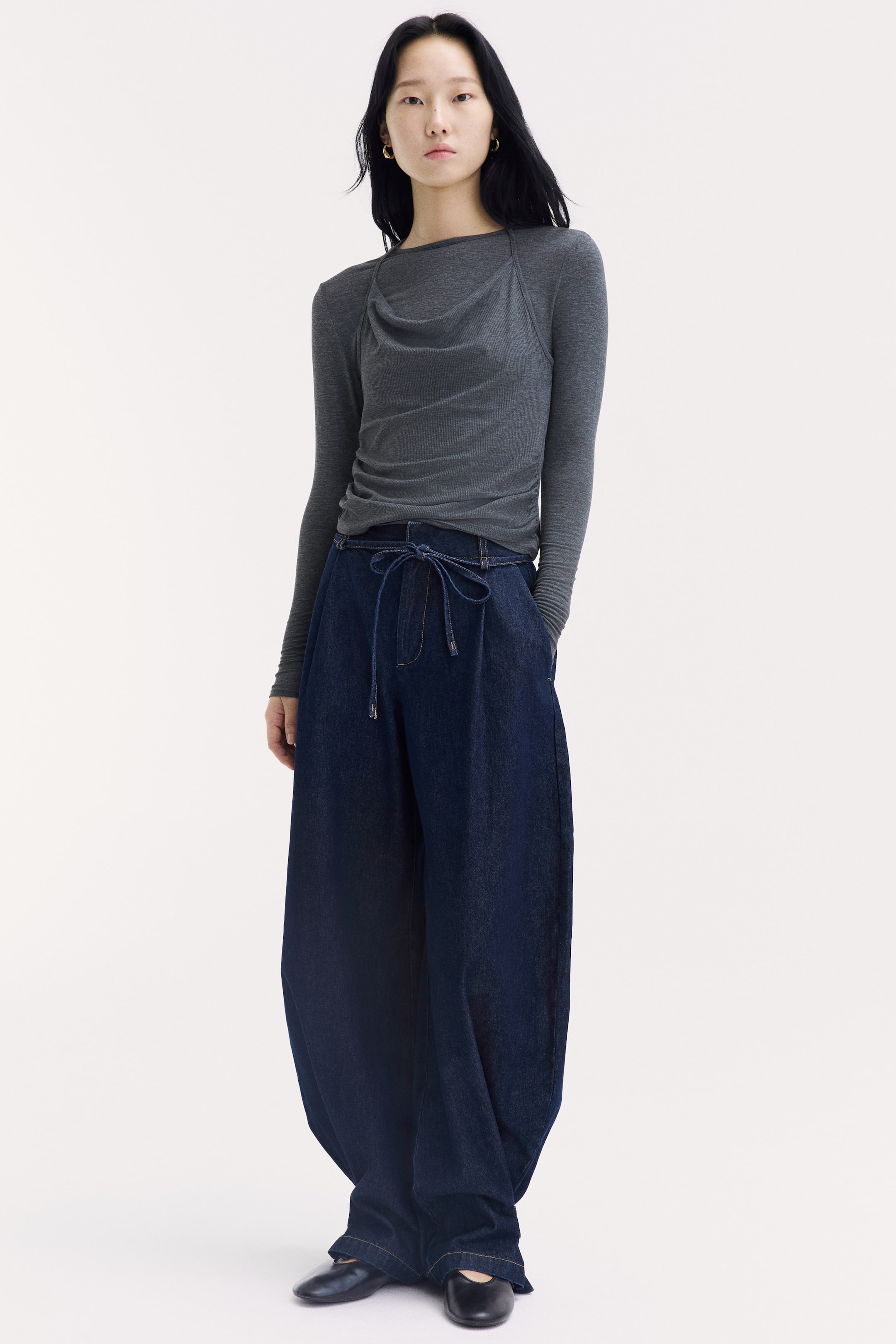 View larger image: The model is wearing a dark grey long-sleeved jersey top, featuring a round neckline with a draped cowl-neck front panel and thin halterneck straps. The top has gathered side seams at the waist and is styled with loose, wide-leg dark denim trousers.