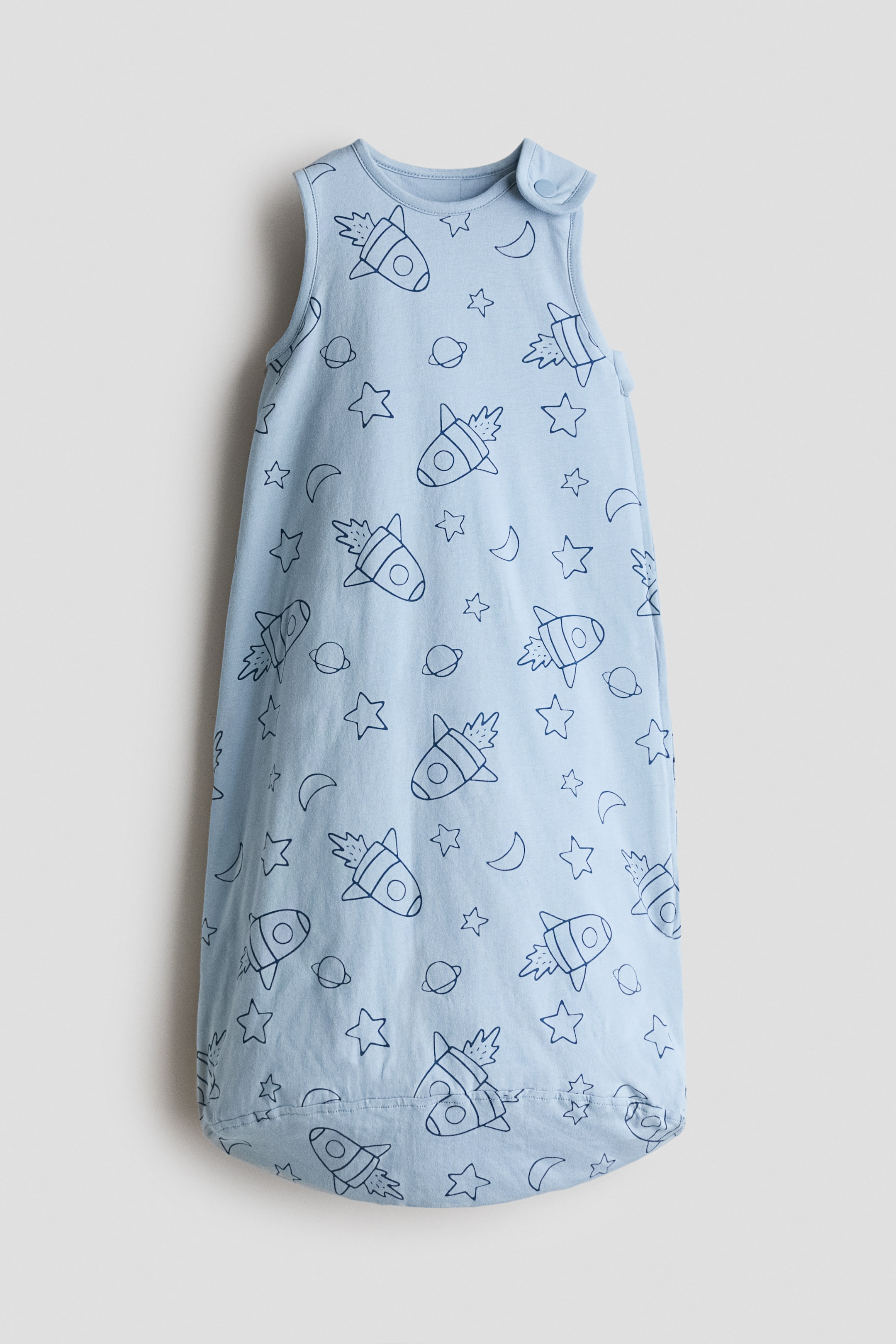 Zobrazit větší obrázek: Polstrovaný spací pytel je prezentován čelním pohledem, vyrobený z lehké modré potištěné bavlněné jersey látky s tmavě modrým vesmírným motivem raket, hvězd a měsíců. Je navržen s patentkovým zapínáním na jednom rameni a zipem, který vede po jedné straně.