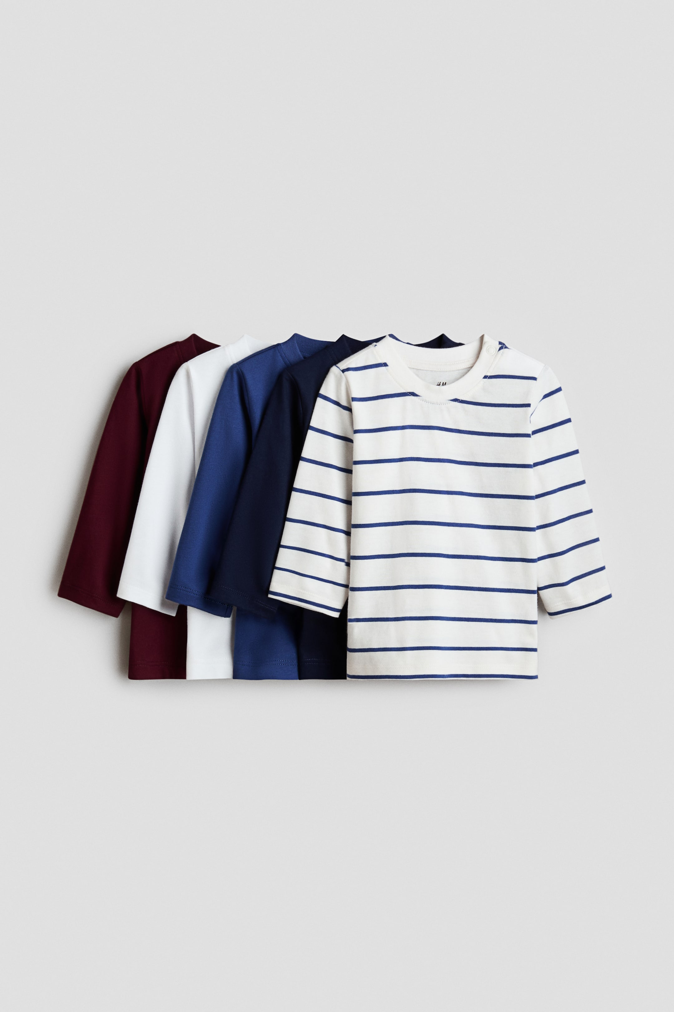 View larger image: Five long-sleeved T-shirts are laid out front-facing in an overlapping arrangement, featuring maroon, white, royal blue, navy blue, and a striped off-white T-shirt with navy blue horizontal lines. The striped T-shirt features two small snap buttons on its left shoulder seam.