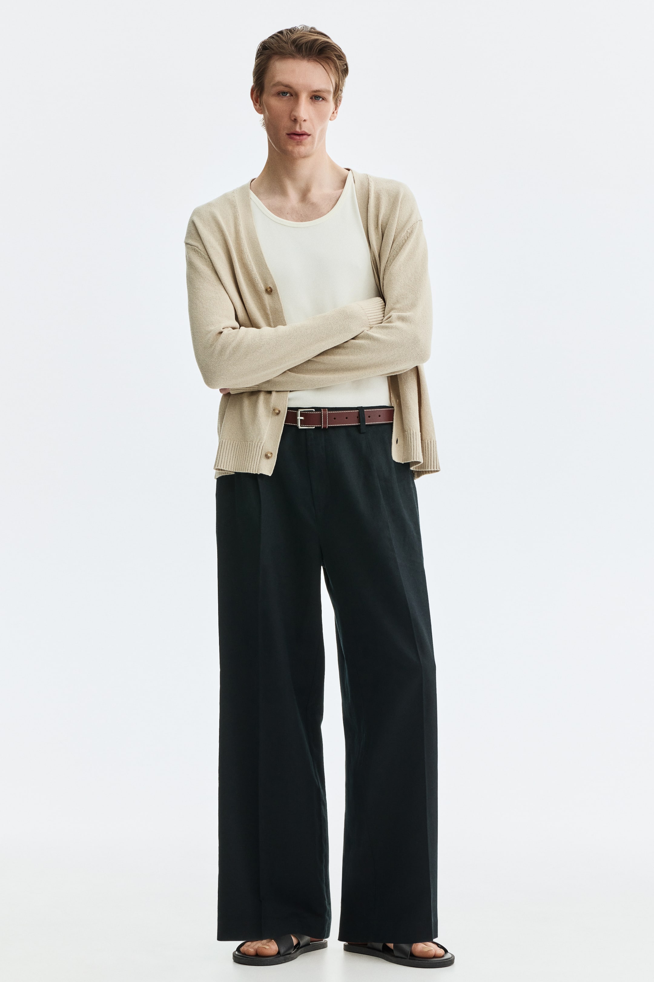 View larger image: Dark tailored trousers, showcasing a relaxed fit, wide legs with pleats at the top, and slant side pockets, are donned by the model. A brown belt is threaded through the waistband, and a zip fly with a button is visible.